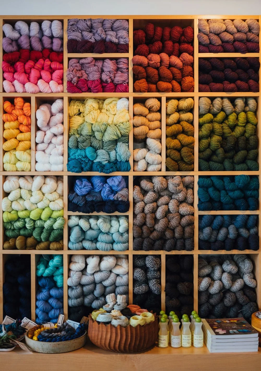 Shelves filled with colorful yarn skeins organized by color, with small bottles and a basket of yarn balls on the table below.