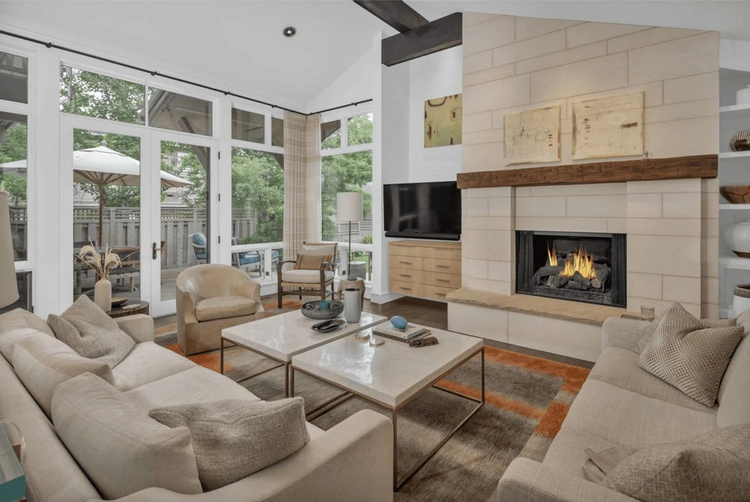 Living room with large windows, fireplace, beige sofas, armchair, coffee table, and patio visible through the windows.