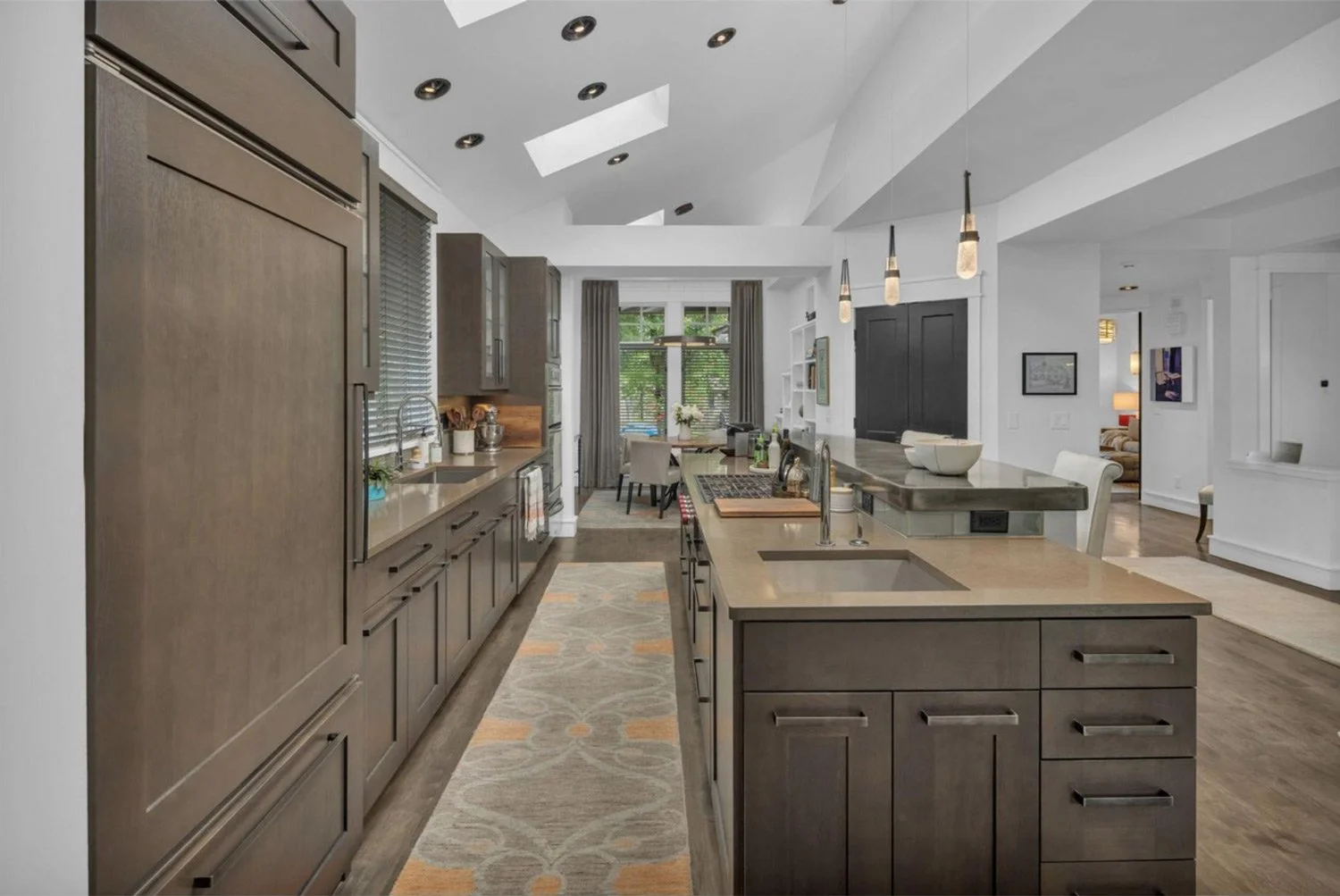 Modern kitchen with gray cabinetry, a large island with a sink, and pendant lights, open to a dining area and living space with large windows and hardwood floors.