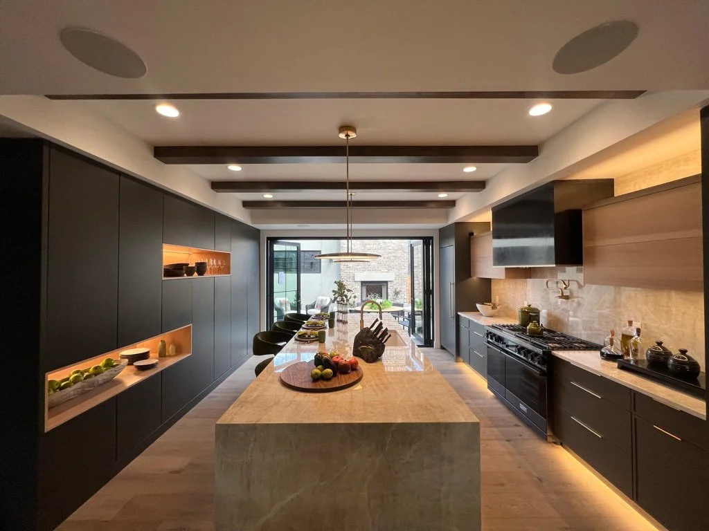 Modern kitchen with a large central island, black cabinets, and built-in shelving with warm lighting, stainless steel appliances, and large glass doors leading outside.