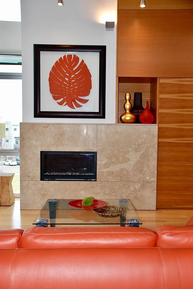 A modern living room with a rectangular fireplace, a framed orange leaf art print above the fireplace, a glass coffee table with a red dish and decorative green moss ball, red and gold vases on a wooden shelf, and a red leather sofa in the foreground
