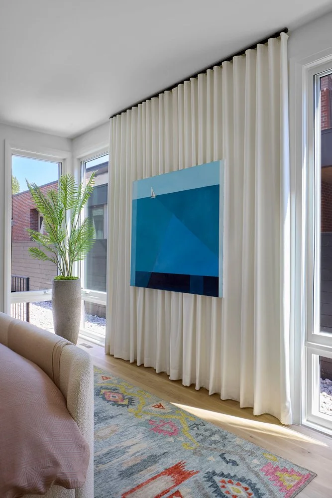 A living room corner with white curtains, a large potted plant, a sofa with pink fabric, a colorful patterned rug, and a blue abstract painting on the wall near windows that let in natural light.