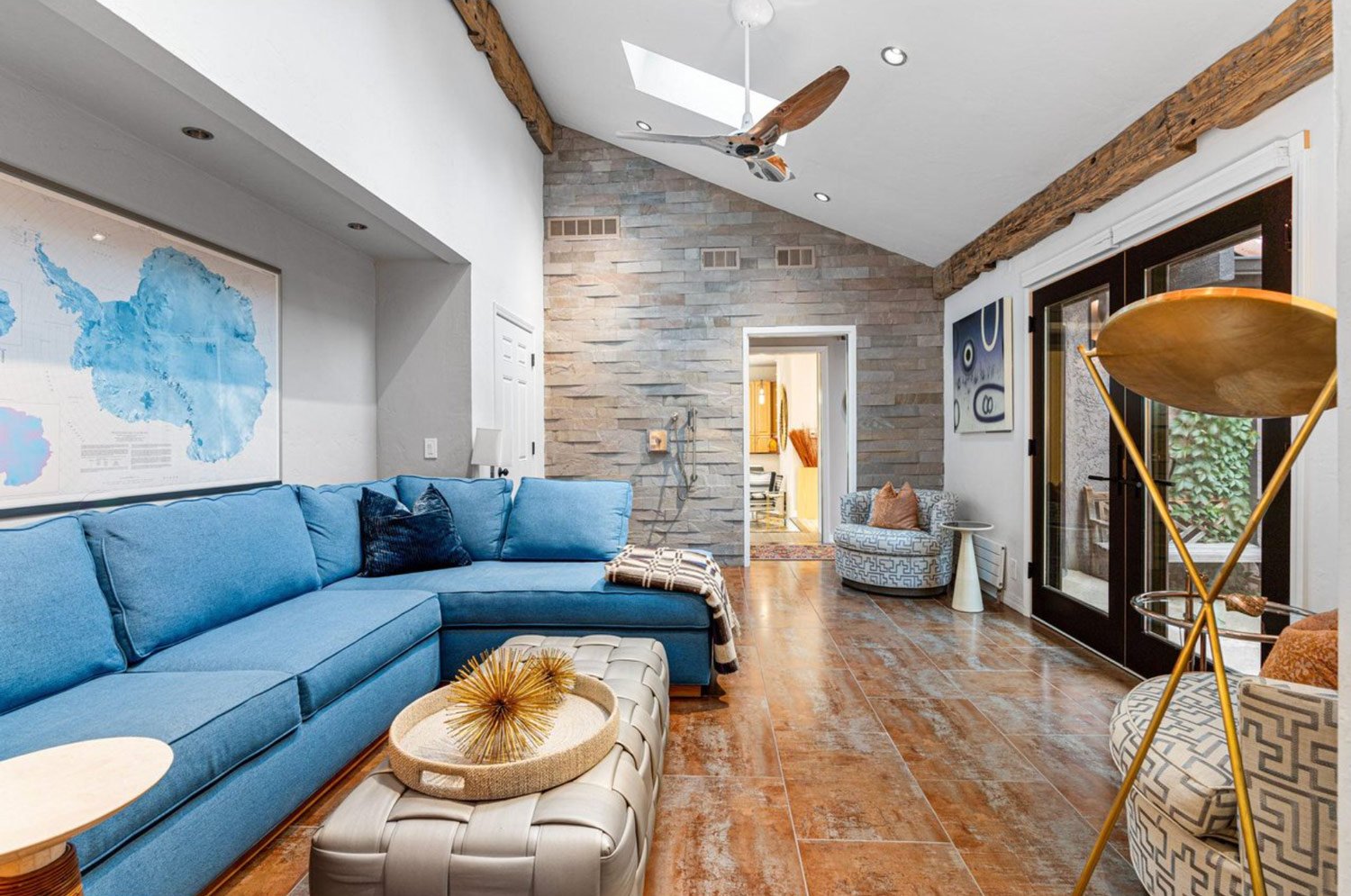 Living room with a large blue sectional sofa, a beige ottoman with a decorative gold spiky orb, a map of Alaska on the wall, a skylight, a modern ceiling fan, a stone accent wall, sliding glass doors, and various chairs and artwork.