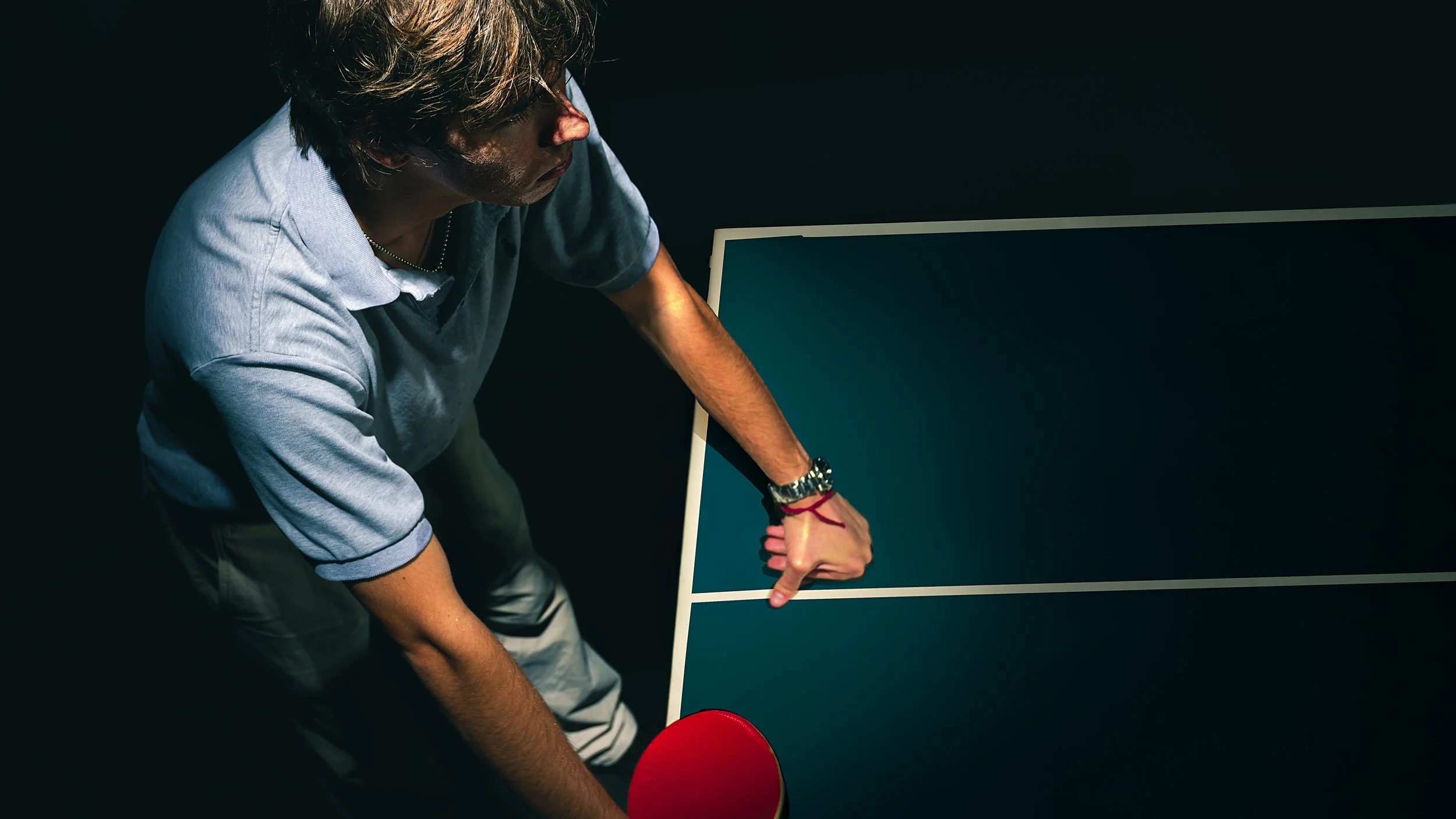 A person with brown hair and glasses leaning over a table with a ping pong paddle and ball, in a dark room with a black background.