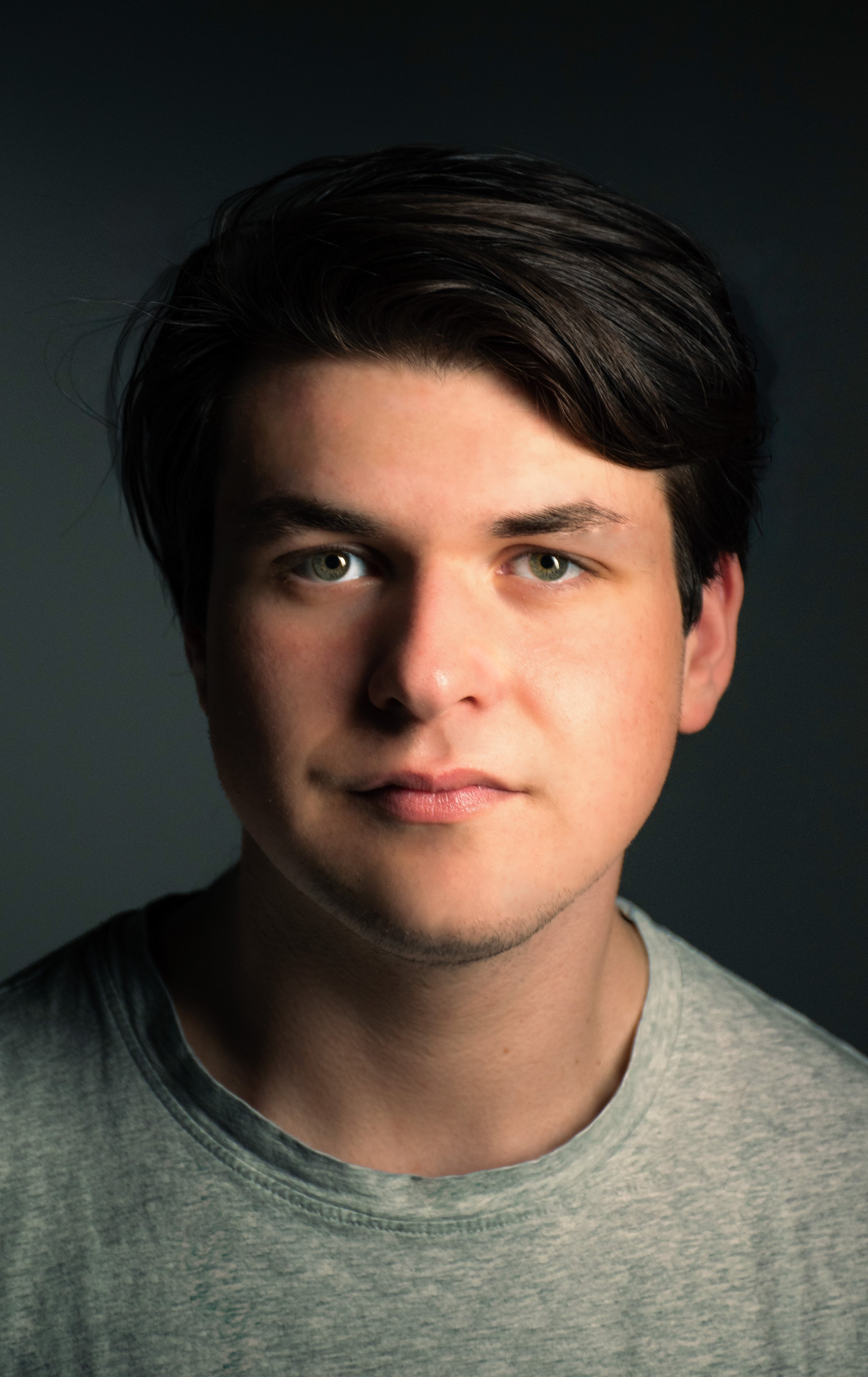 Close-up of a young man with dark hair, fair skin, and light-colored eyes, wearing a gray t-shirt, against a dark background.