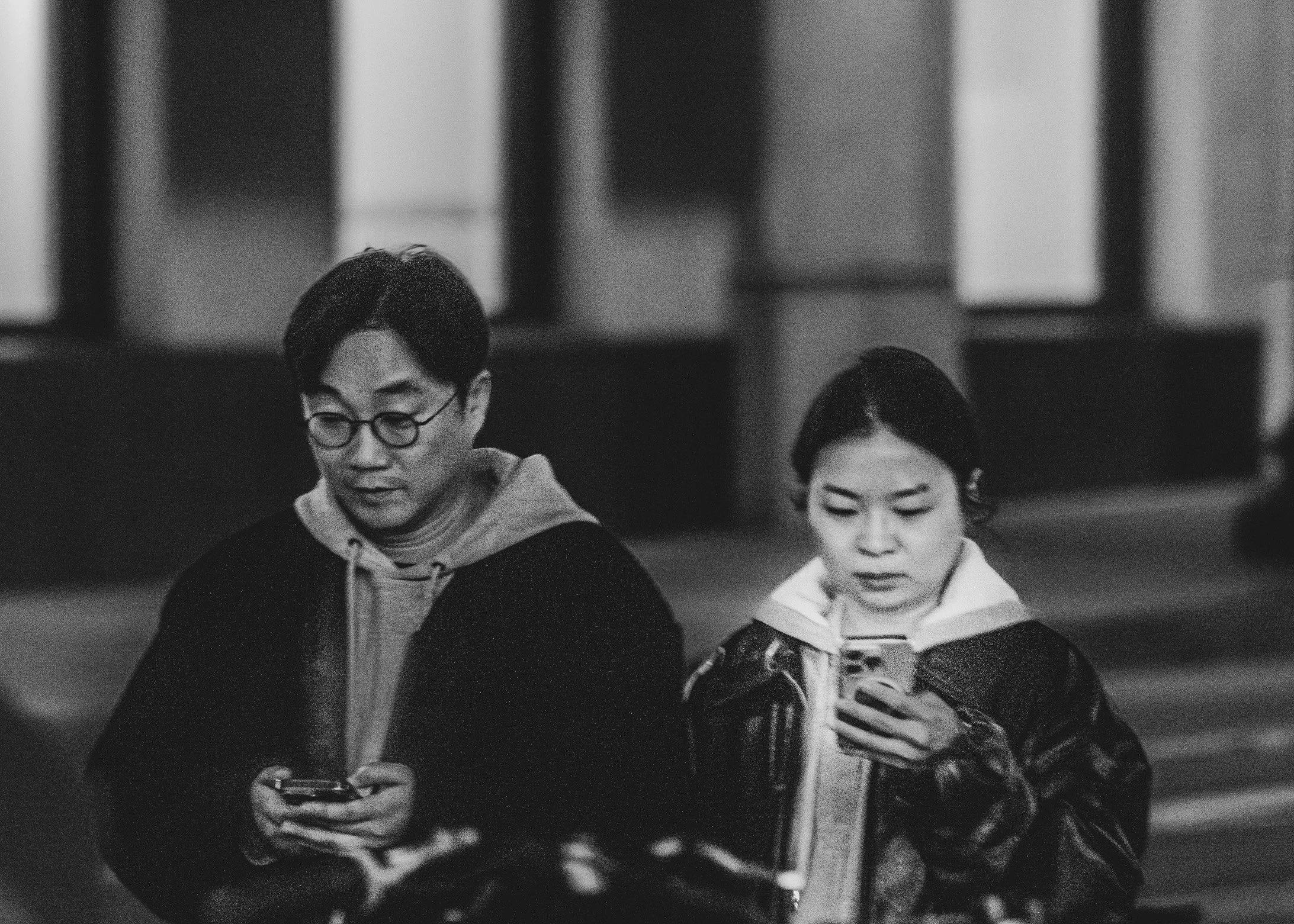 Black and white photo of two people using smartphones, walking indoors with blurred background.