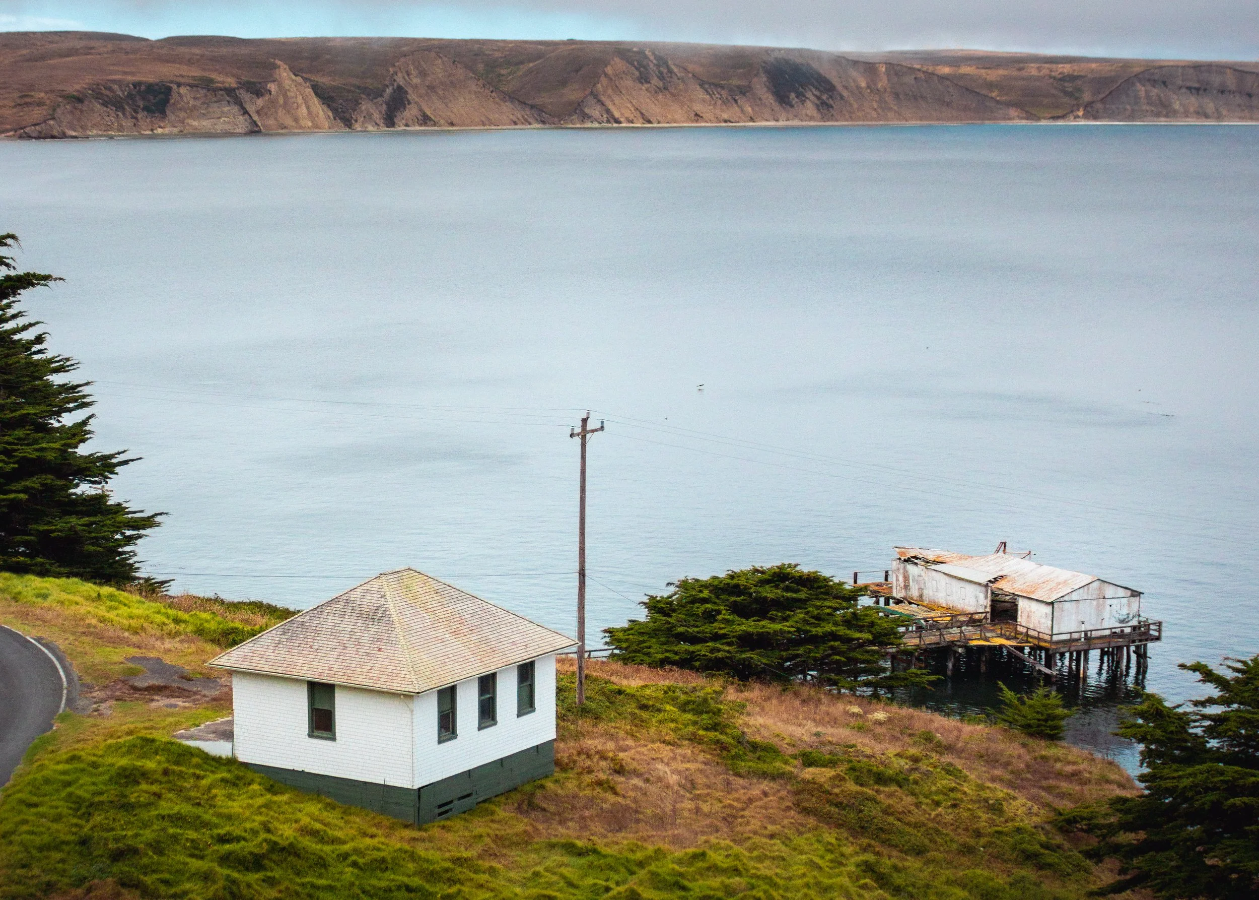 A coastal scene featuring a white house on a grassy hill, with trees and buildings nearby, overlooking a calm body of water with brown rocky cliffs in the background.