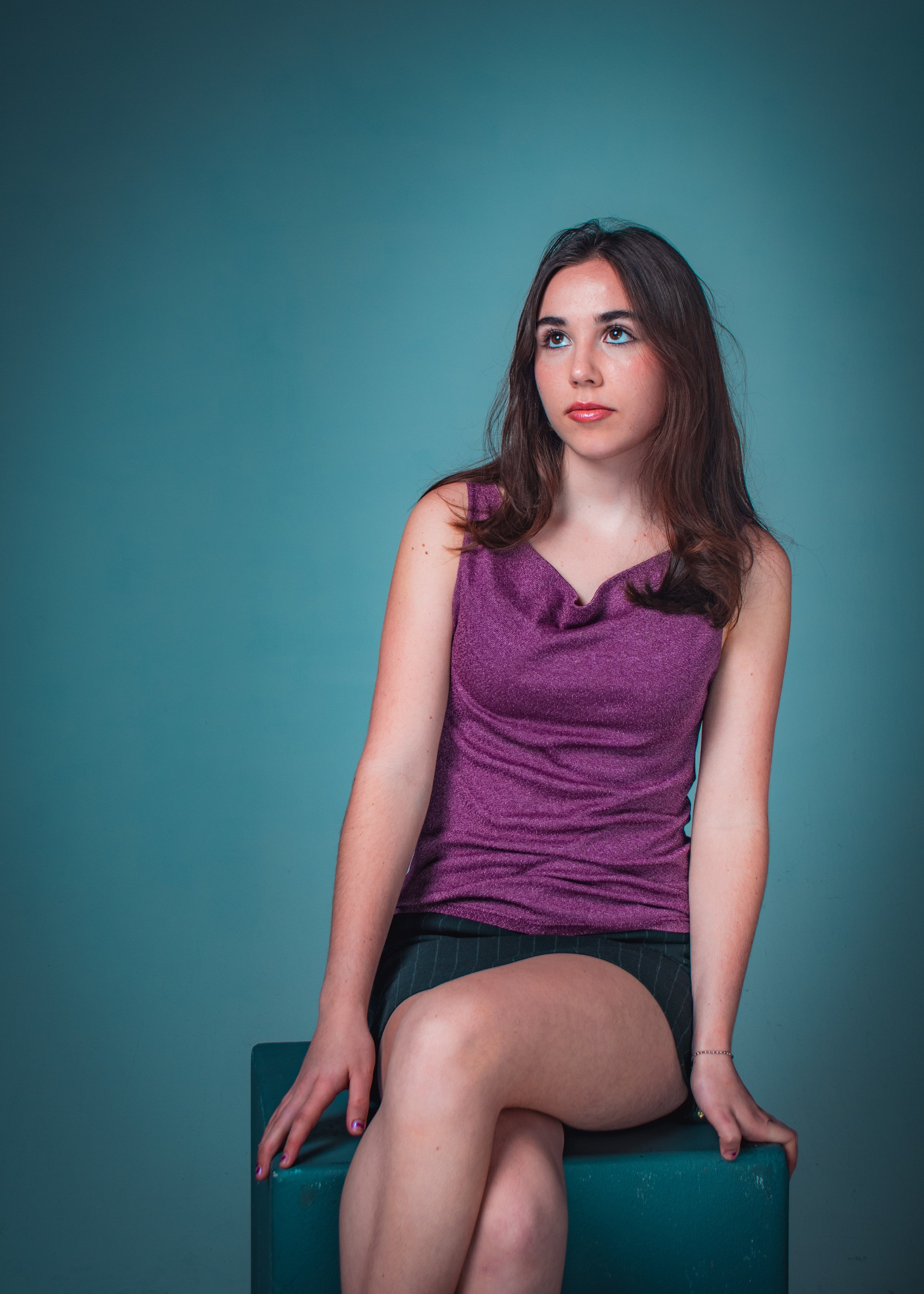 A young woman with brown hair wearing a purple sleeveless top and black shorts sitting on a teal chair against a teal background, with her right leg crossed over her left knee and her hands resting on her lap.