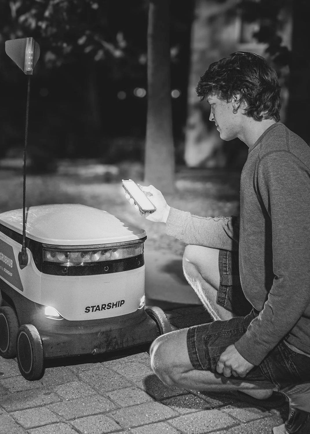 A person kneeling on the sidewalk at night, looking at a smartphone, with a small robotic vehicle labeled 'STARSHIP' nearby, and an arrow sign to the left.