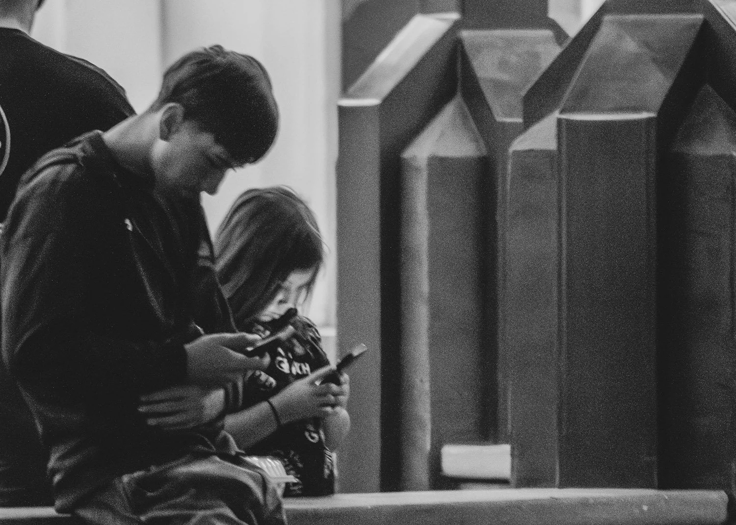 Two teenagers sitting on a bench, looking at their smartphones, with a third person partially visible beside them, in black and white.