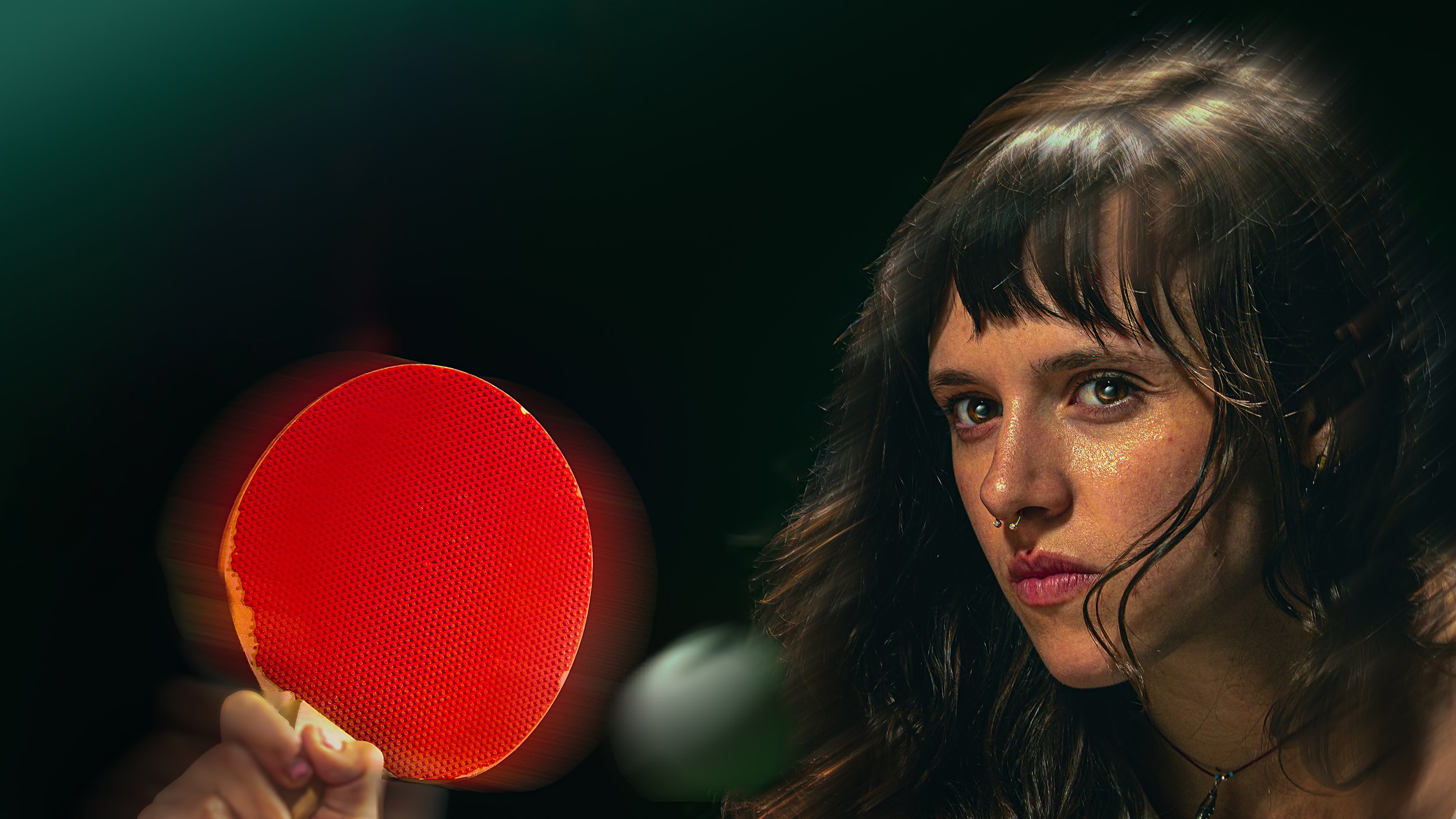 A woman holding a red ping pong paddle near her face, with dark hair and intense expression, in a dark background.