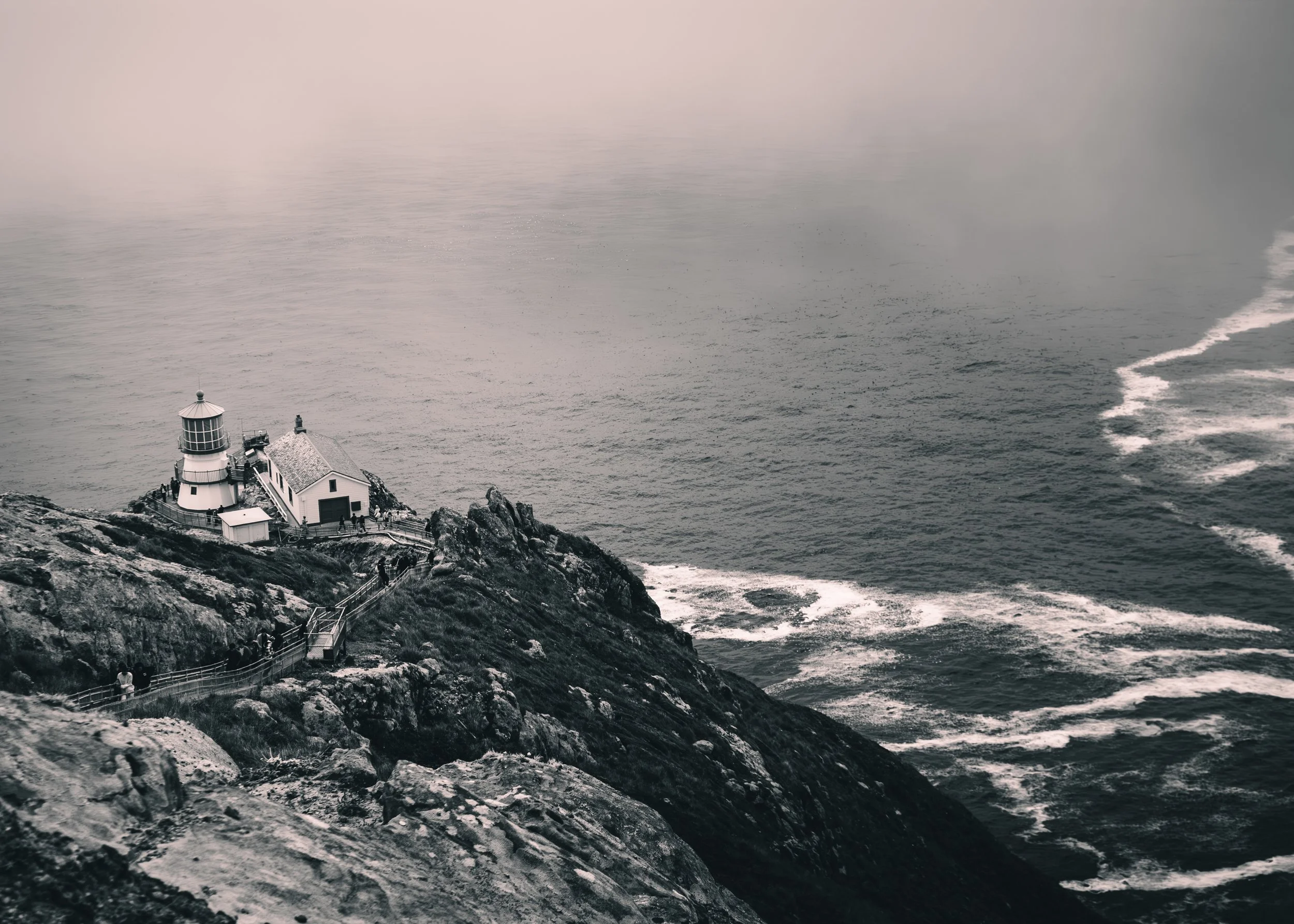 A lighthouse on a rocky coastline with a walkway and several visitors, during foggy weather over the ocean.