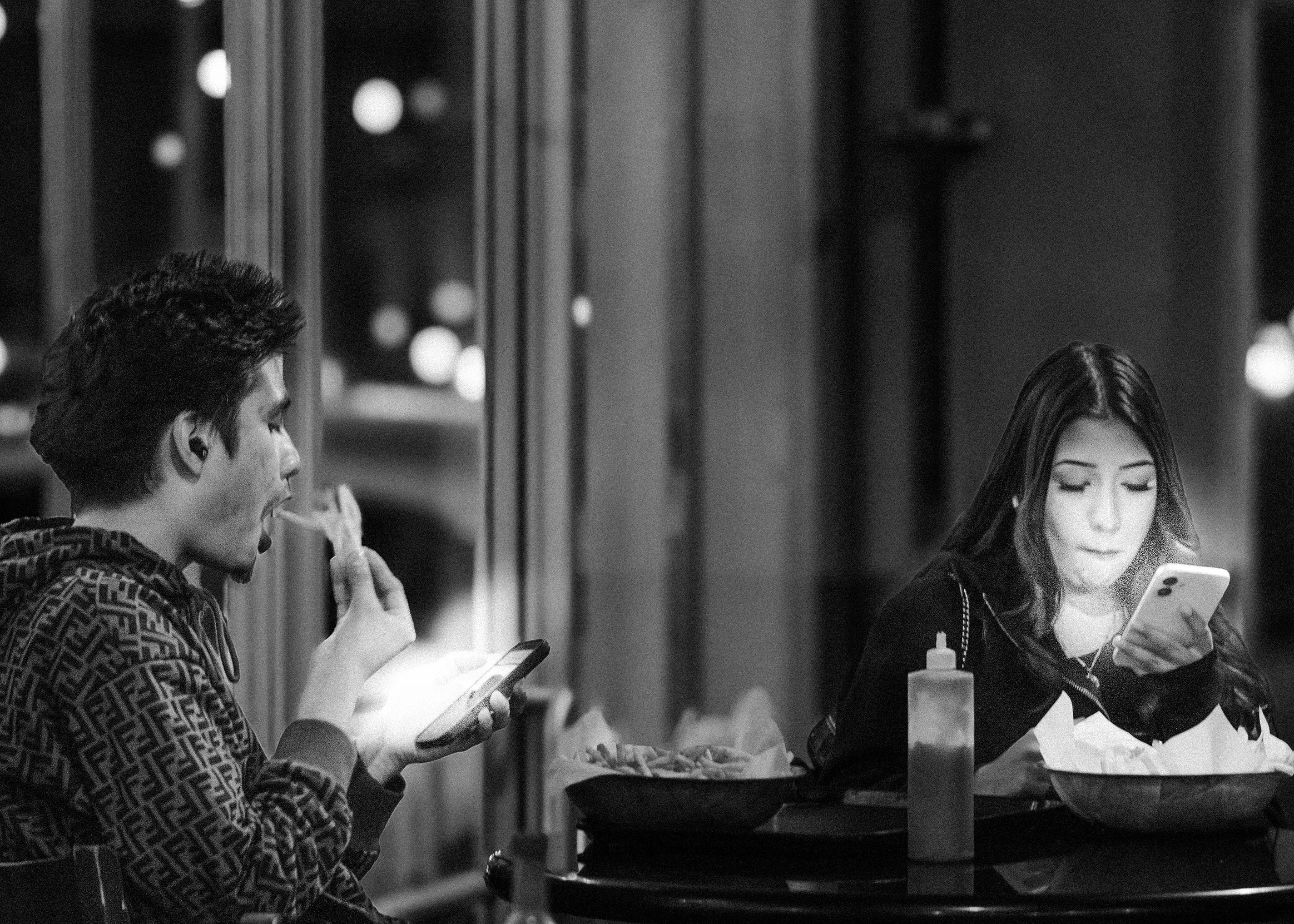 Two people sitting at an outdoor table, looking at their phones. The man on the left is drinking from a glass with a straw, and the woman on the right is holding a phone and looking at it. There is a large bucket with bottles on the table.