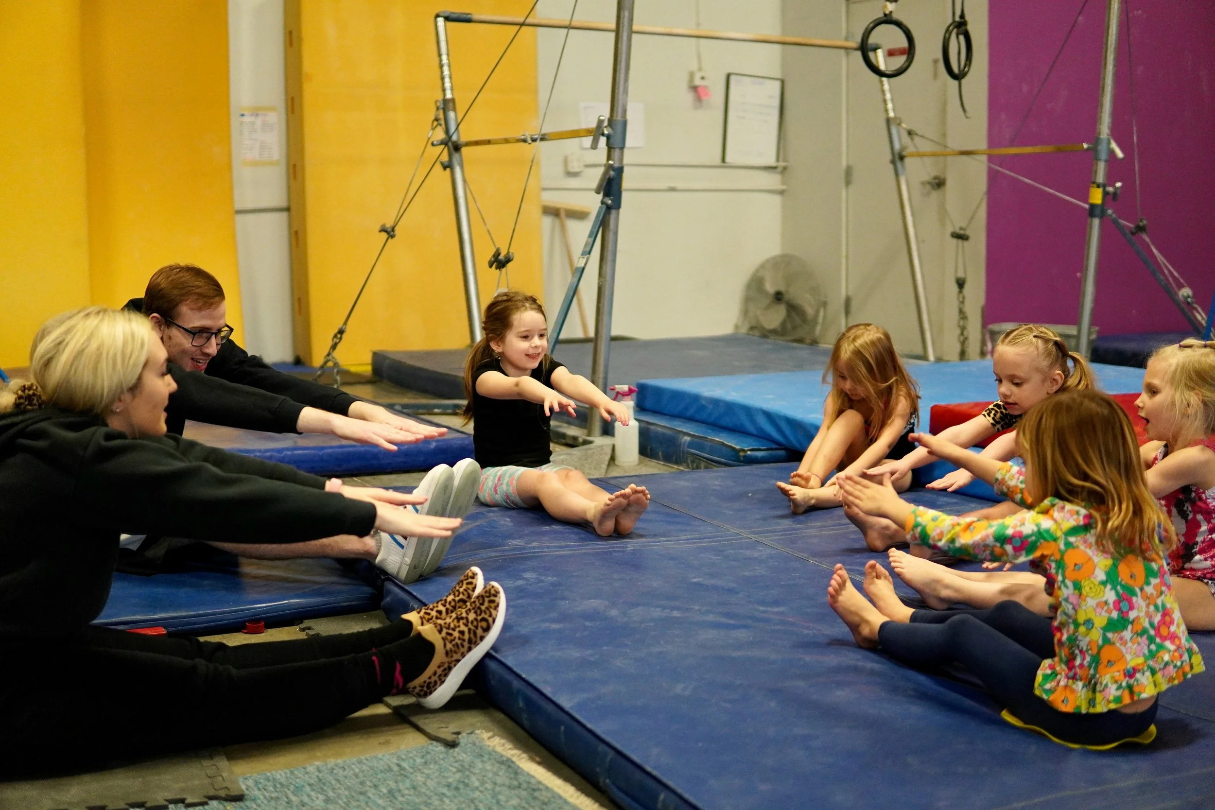 Children and adults sitting on a blue mat in a gym, participating in a stretching exercise with legs extended and reaching for their toes. Gymnastic equipment like rings and a climbing frame are visible in the background.