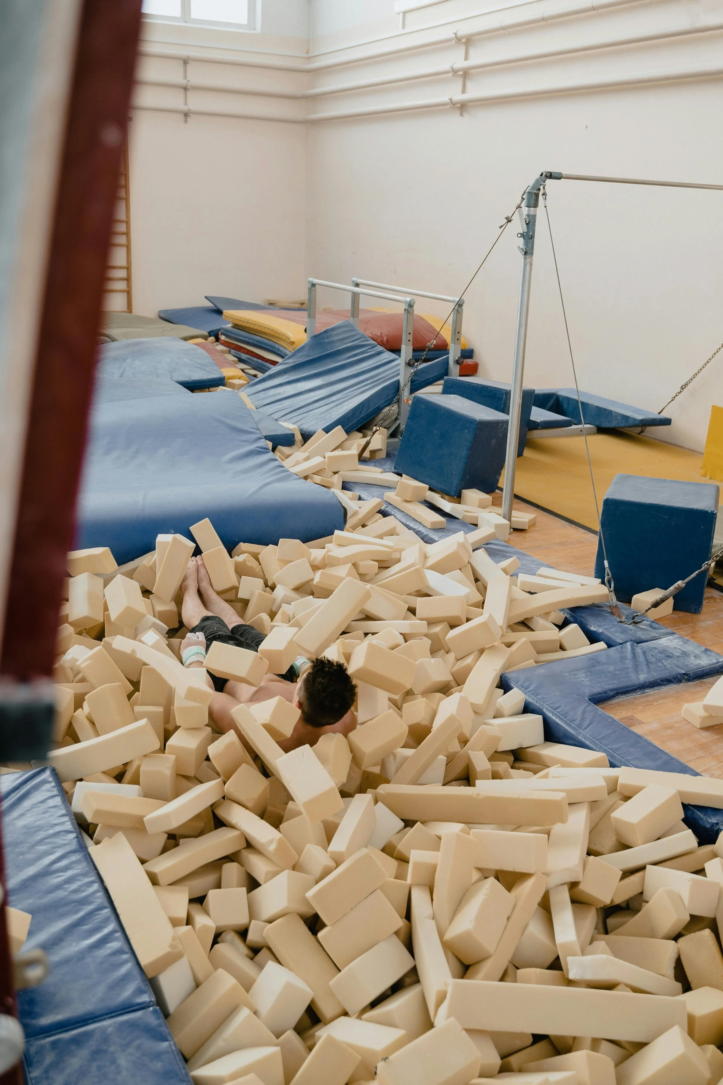 Indoor gym with foam pit filled with beige foam blocks, foam climbing apparatus with blue and gray padding, some stacked mats, and a person lying in the foam.