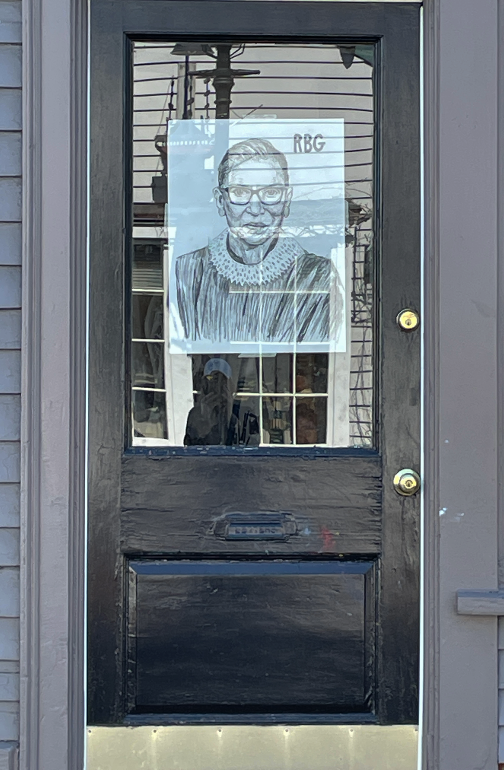 Reflection of electrical wires and an outdoor landscape seen through a glass door, with a portrait of a woman wearing glasses and a top with a lace collar taped to the door.