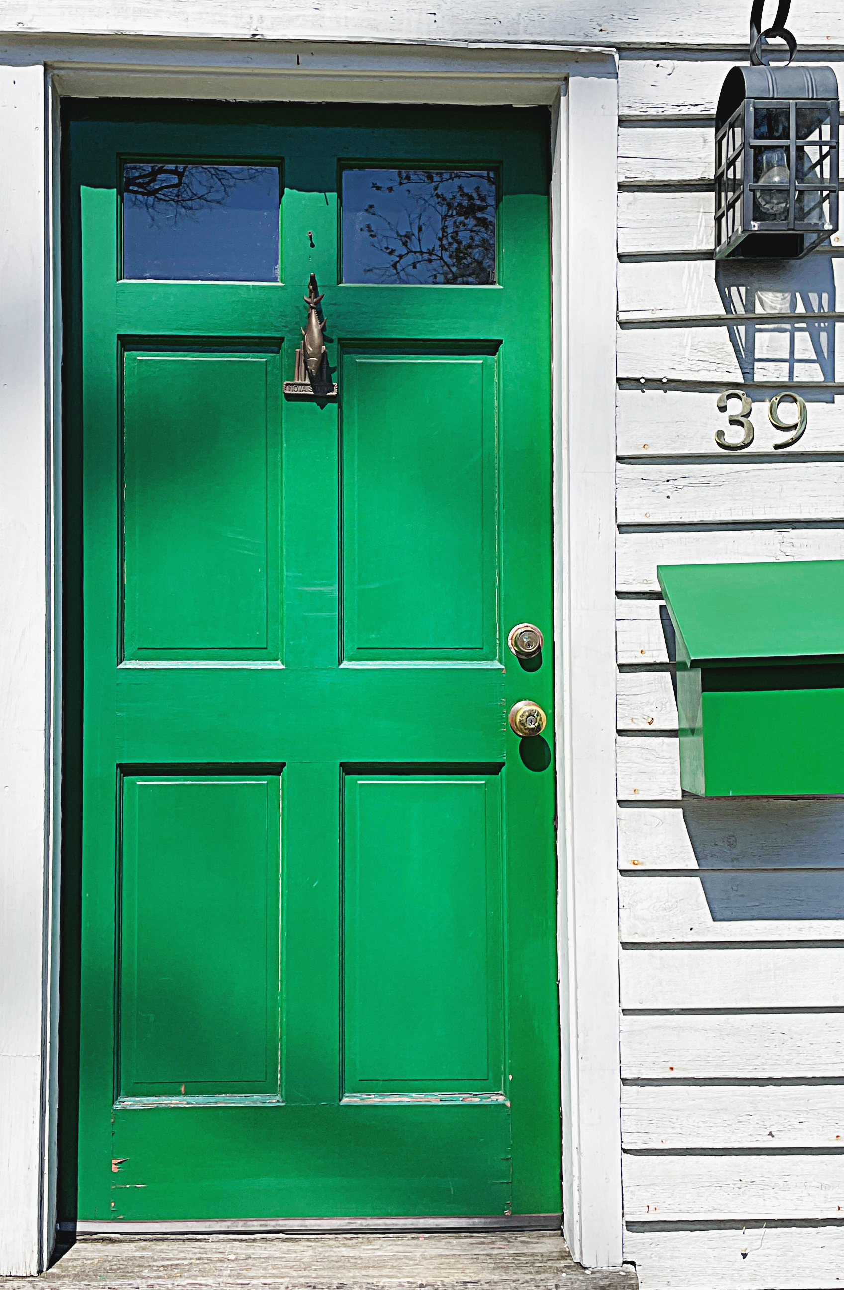 Green front door with a window at the top, a fish-shaped door knocker, and house number 39 on the white wooden house siding. A black outdoor lantern is mounted on the wall to the right of the door, and a green mailbox is below it.