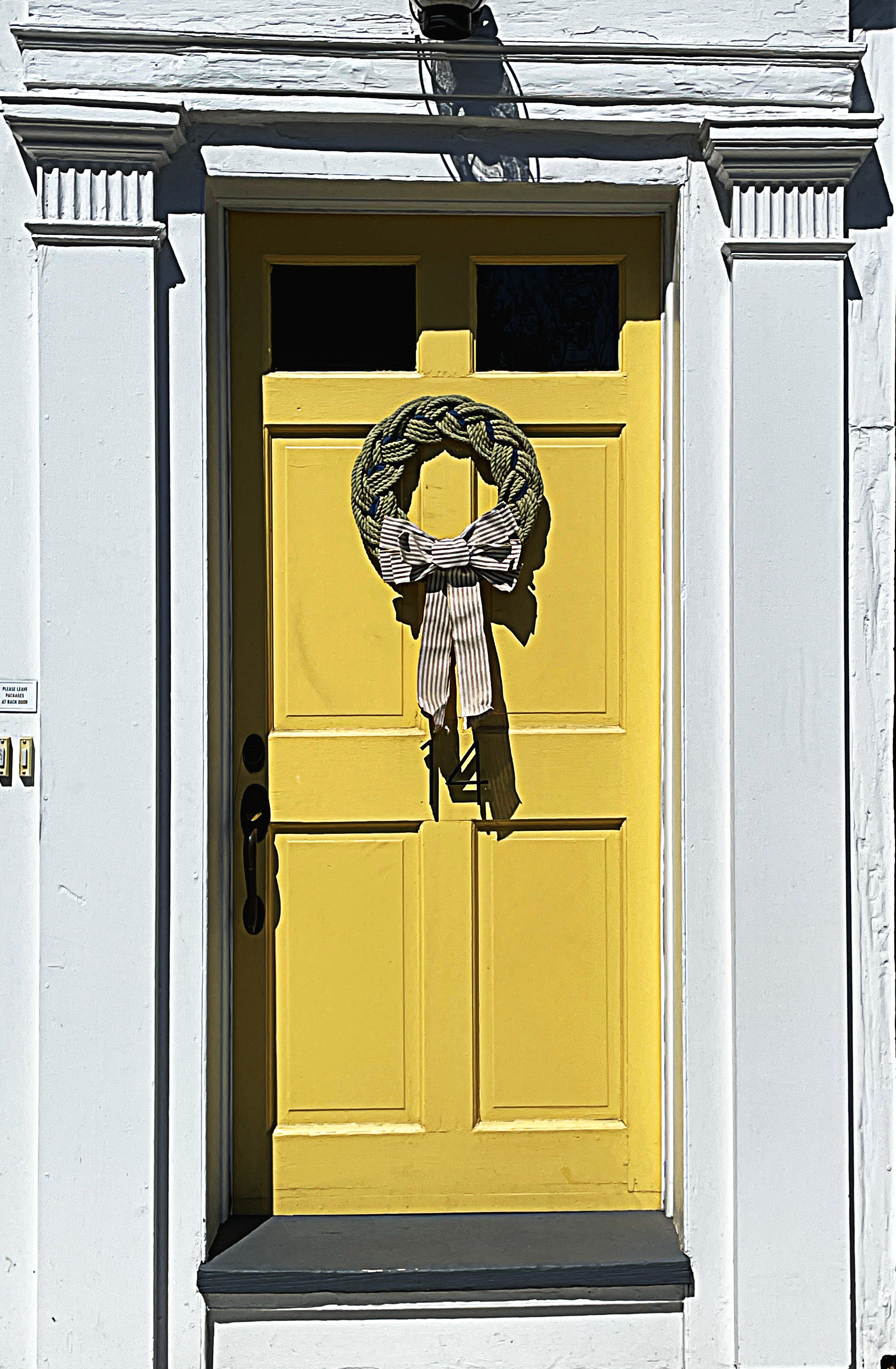 Close-up of a yellow door with a decorative wreath with a striped ribbon in the center, set in a white wall with architectural molding. The wreath is made of braided rope and the door has a small window at the top.