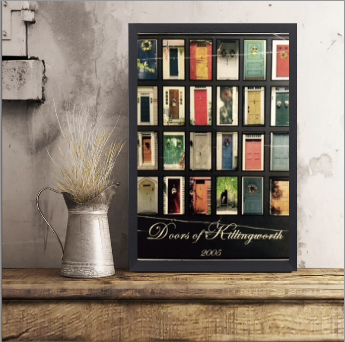 A vintage silver pitcher with dried white and beige grasses, placed on a wooden surface next to a framed collage of colorful doors labeled "Doors of Killington, 2005."