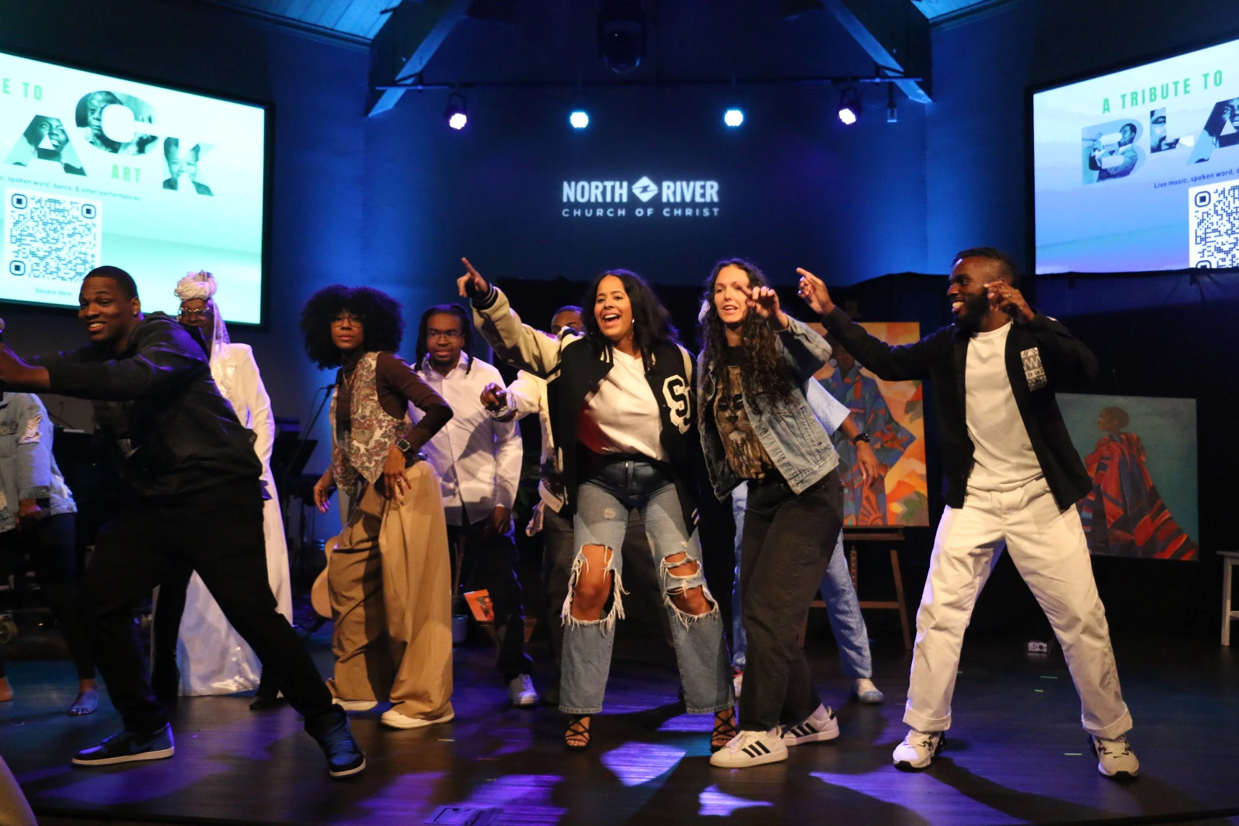 Group of diverse people dancing and singing on stage at North River Church of Christ, with two large screens displaying a tribute to Black art and culture in the background.