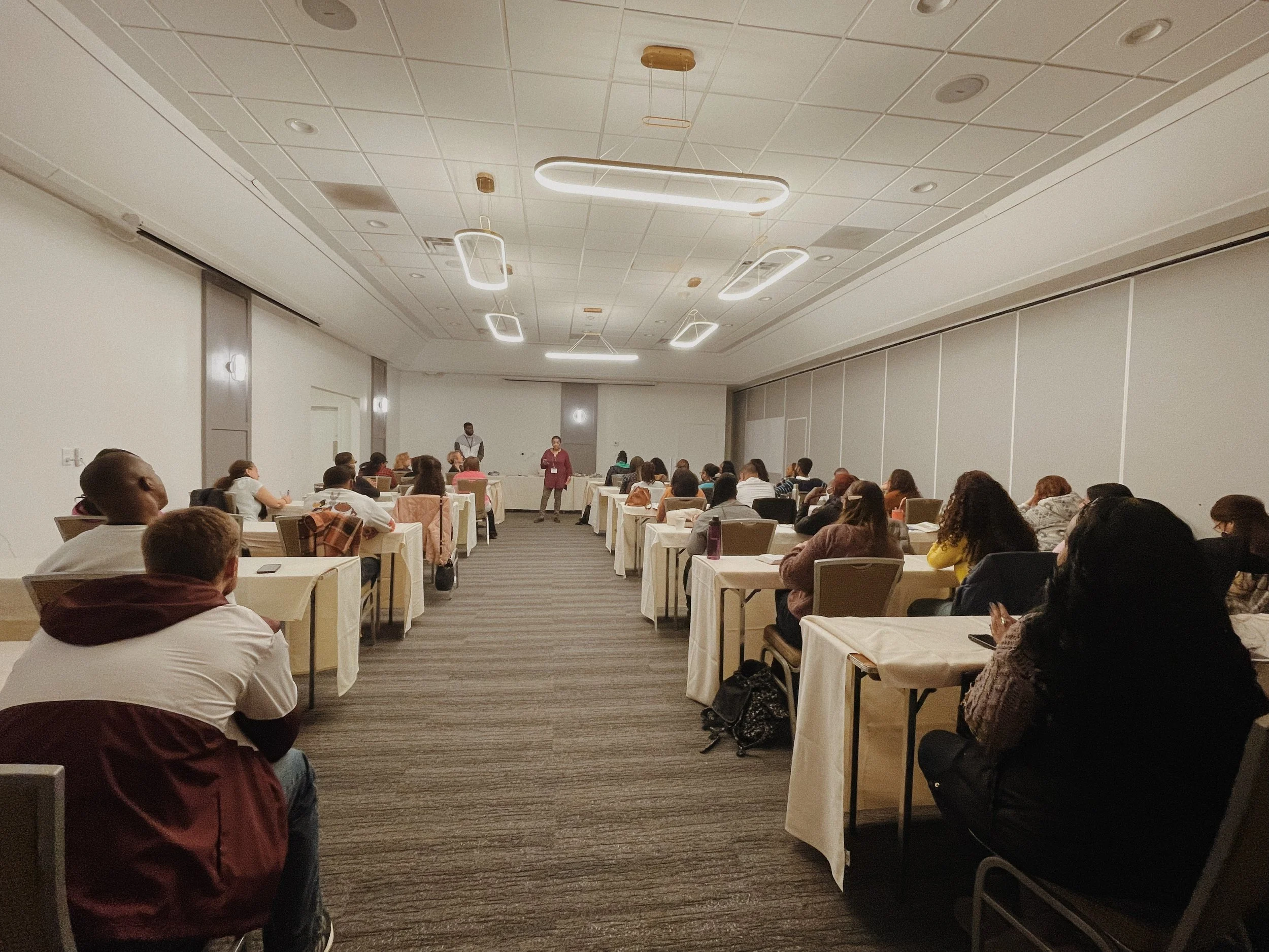A large conference room filled with people sitting at tables, facing a speaker standing at a podium. The room has a high ceiling with modern rectangular light fixtures and white walls.
