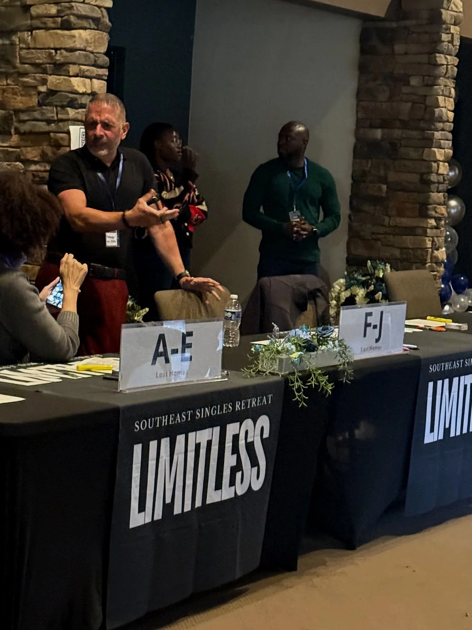 People standing behind a table with signs reading 'Southeast Singles Retreat'. The table is decorated with flowers and balloons. The background has a brick wall.
