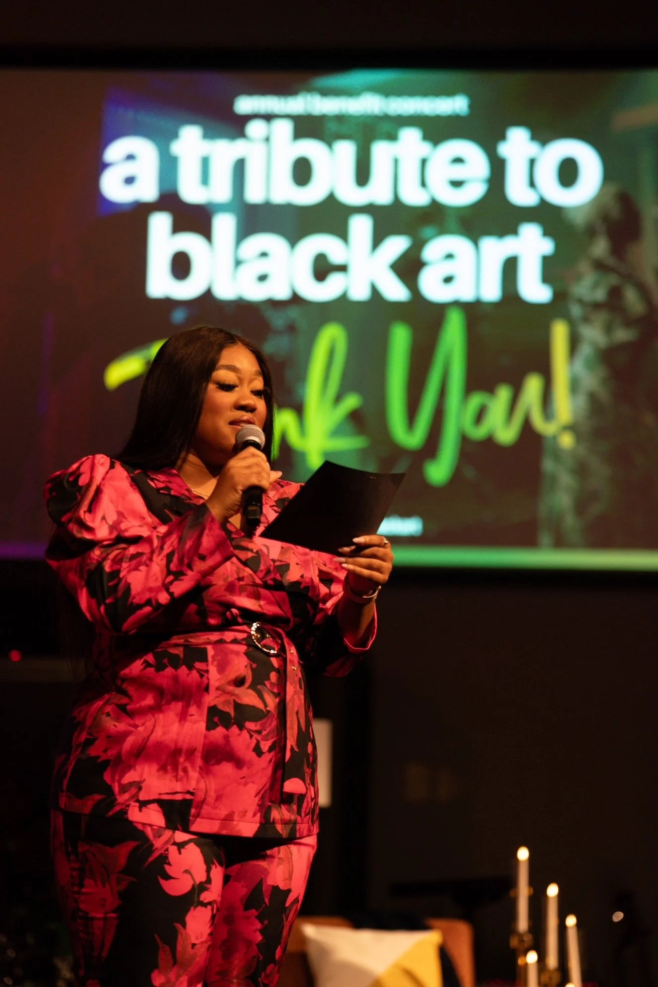 A woman with long black hair wearing a red and black floral outfit is speaking into a microphone on stage. Behind her, a projected slide reads 'a tribute to black art' in white and green letters.