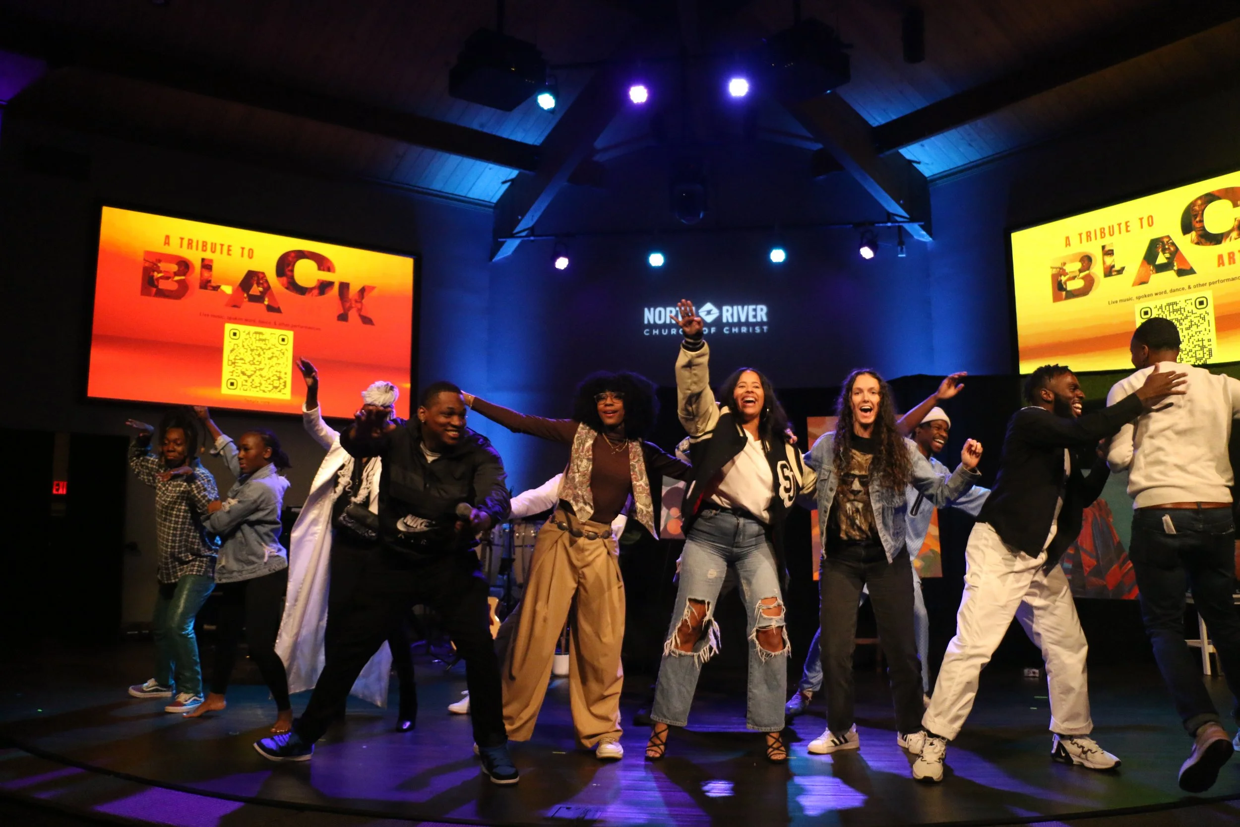 Group of people dancing and celebrating on stage during a live event at North River Church of Christ, with two large screens displaying a tribute to Black culture in the background.