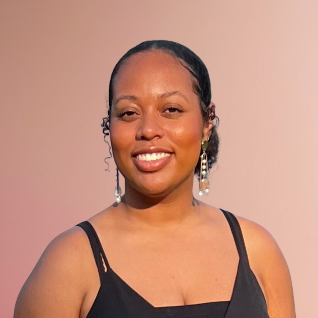 A smiling woman with dark hair styled in a sleek manner, wearing a black spaghetti strap top and dangling earrings, standing against a plain background.