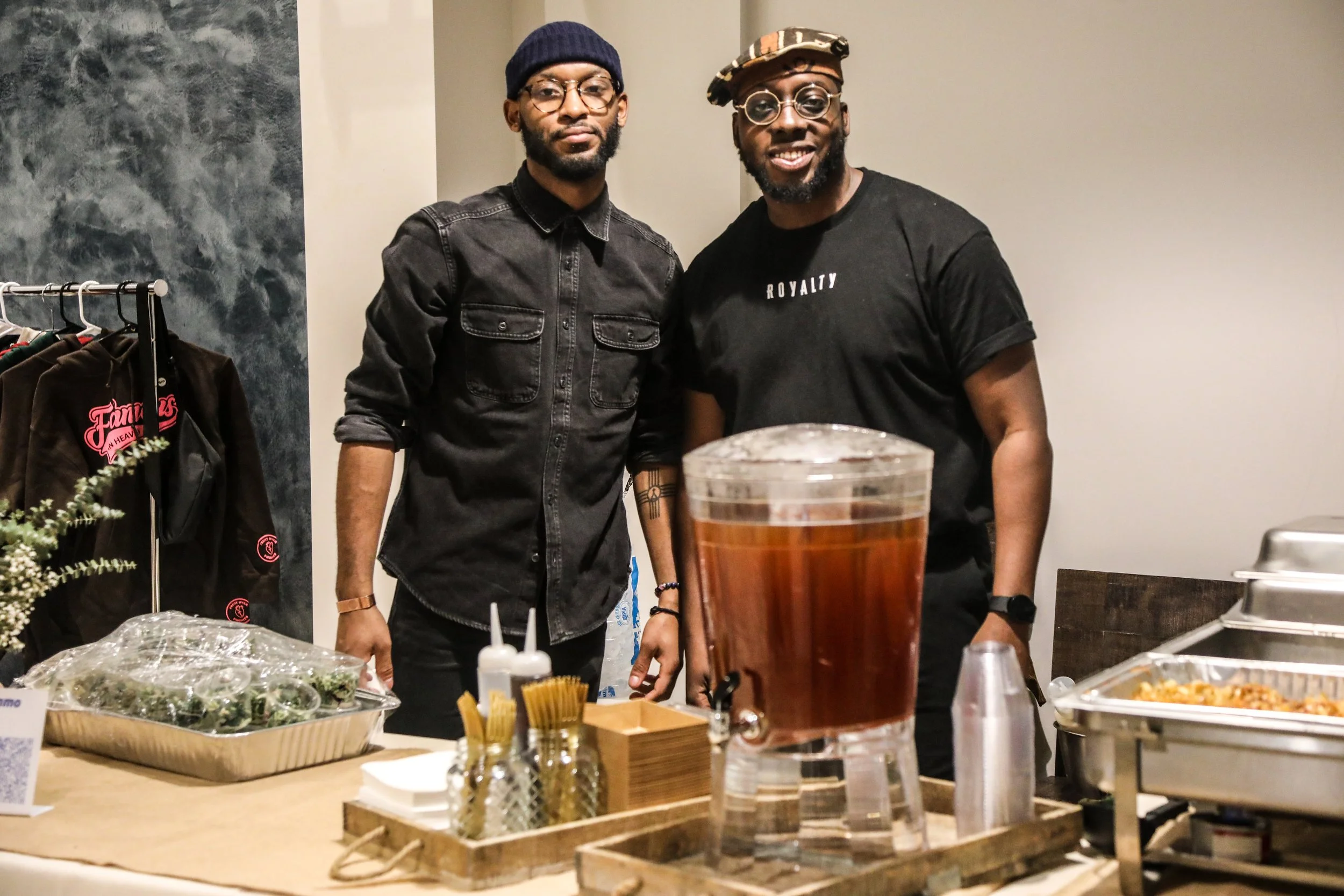Two men standing behind a table with food and drinks, smiling at the camera. One wears a black shirt with 'ROYALTY' written on it and a patterned cap, the other a black denim shirt and a beanie. There is a large glass of iced tea or soda on the table.