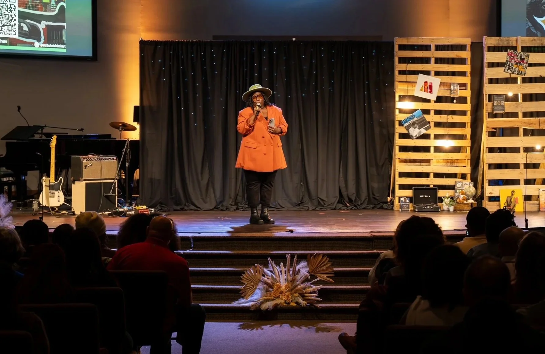 A woman stands on stage, speaking into a microphone in front of a black curtain backdrop with small white lights. She wears an orange blazer, black pants, black boots, and a wide-brimmed hat, and holds a phone. There are musical instruments and equipment to her left, including a guitar and amplifier. To her right, there are wooden pallet dividers with pictures and small objects attached. The audience is seated in front of the stage, with some flowers visible on the floor.
