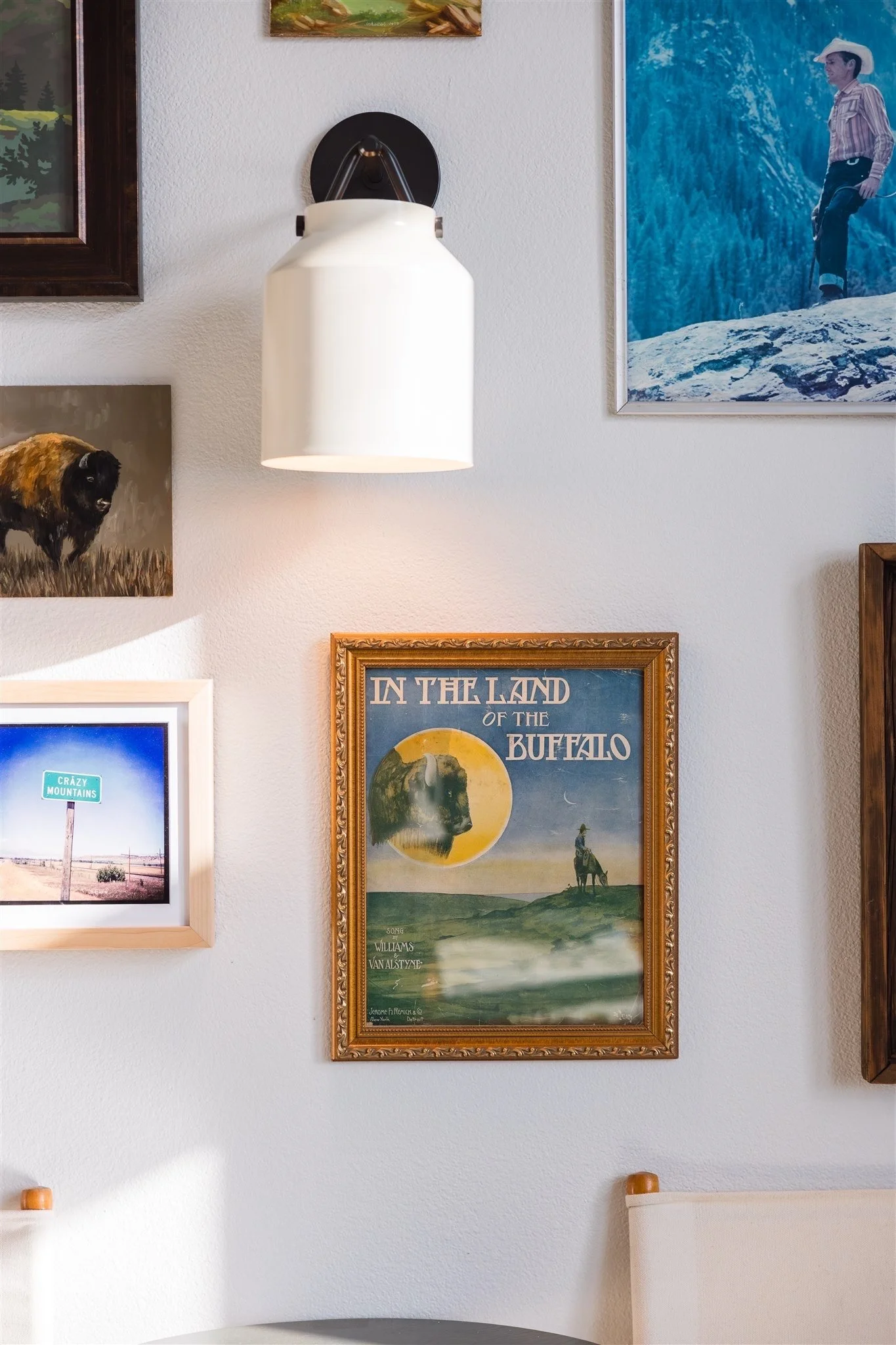 Close-up of a wall decorated with framed pictures and a white . The central framed poster features a cowboy riding a horse, with the text "In the Land of the Buffalo." Surrounding art includes a painting of a mountain landscape, an image of a black b