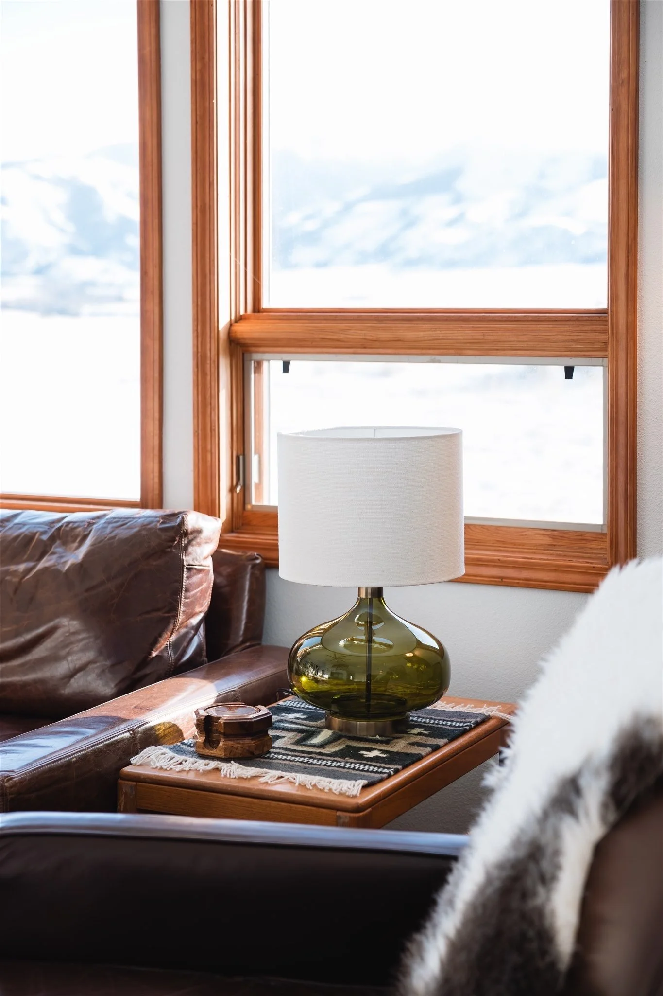 Close-up of a living room corner with a brown leather sofa, a wooden side table with a lamp, and a window with wooden frames showing a snowy landscape outside.