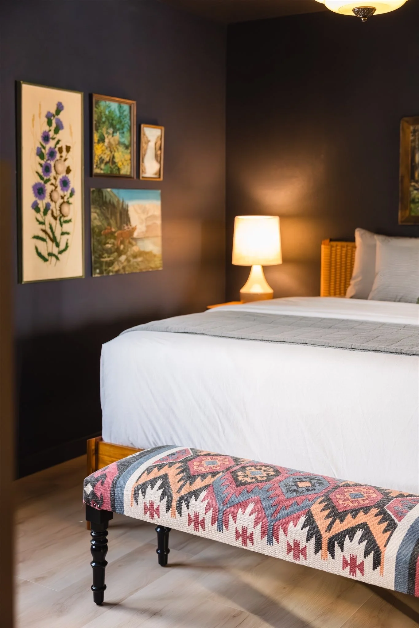 A hotel room with a bed, a nightstand with a lamp, wall art, and a bench with a patterned fabric. The walls are dark-colored, and the floor is wooden.