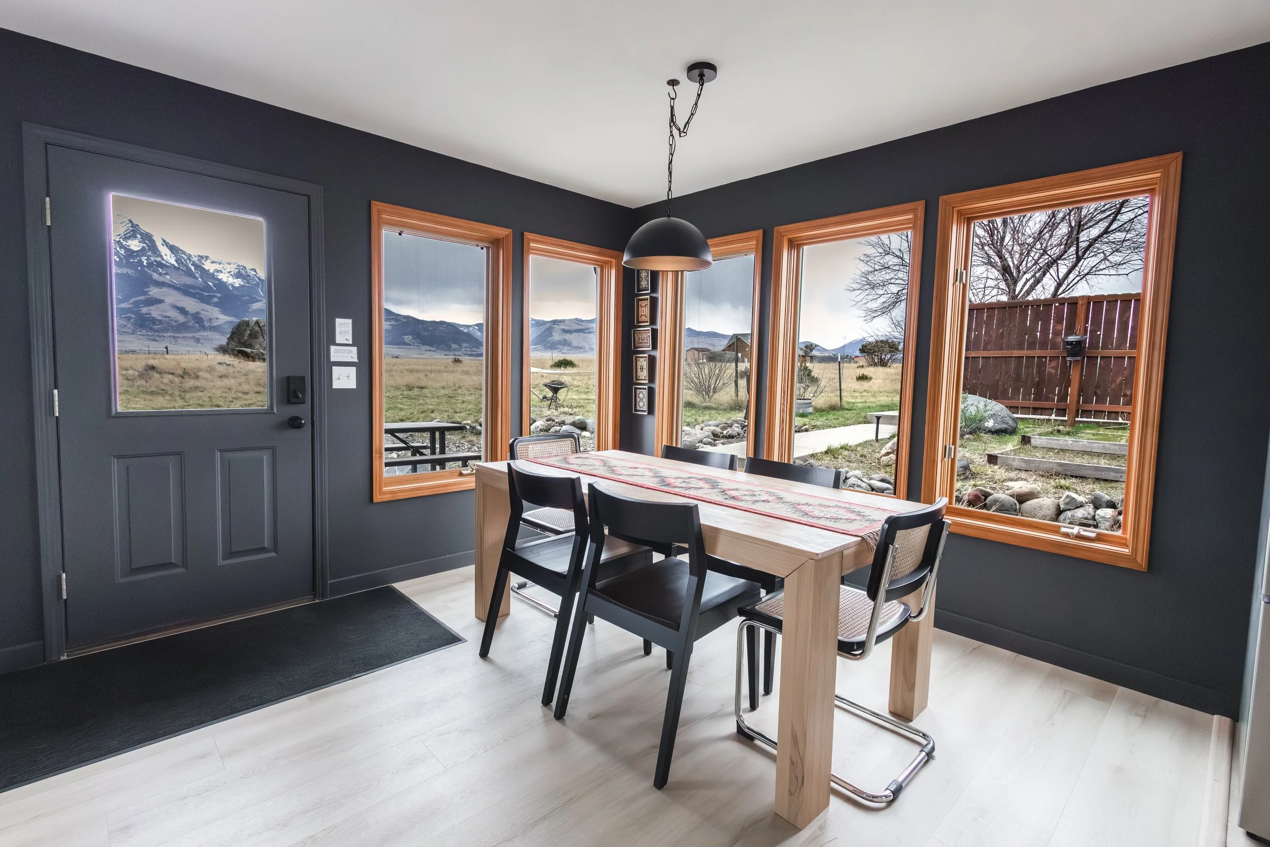Interior of a dining room with four black chairs around a wooden table with a decorative runner, large windows showing mountains and open land outside, and a black pendant light hanging from the ceiling.