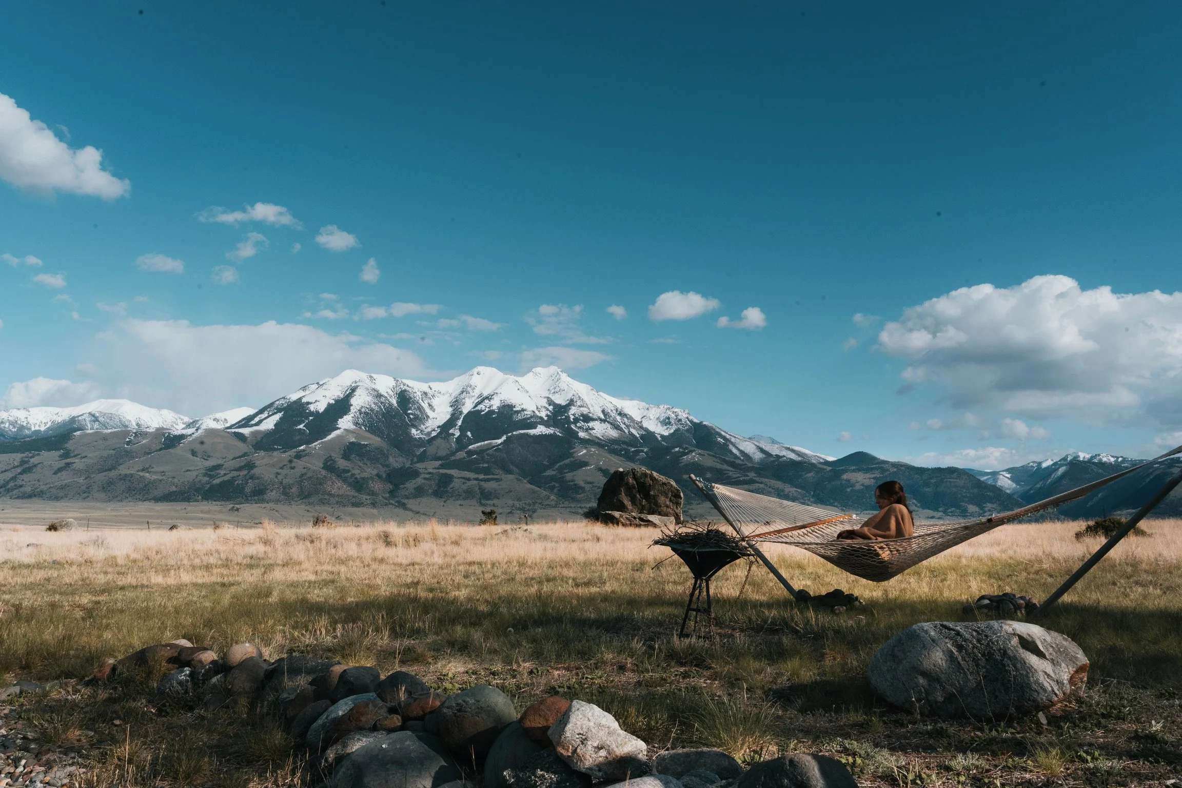 Person relaxing on a Pawleys Island hammock in a backyard field with Emigrant Peak and snow-capped peaks under a partly cloudy sky.
