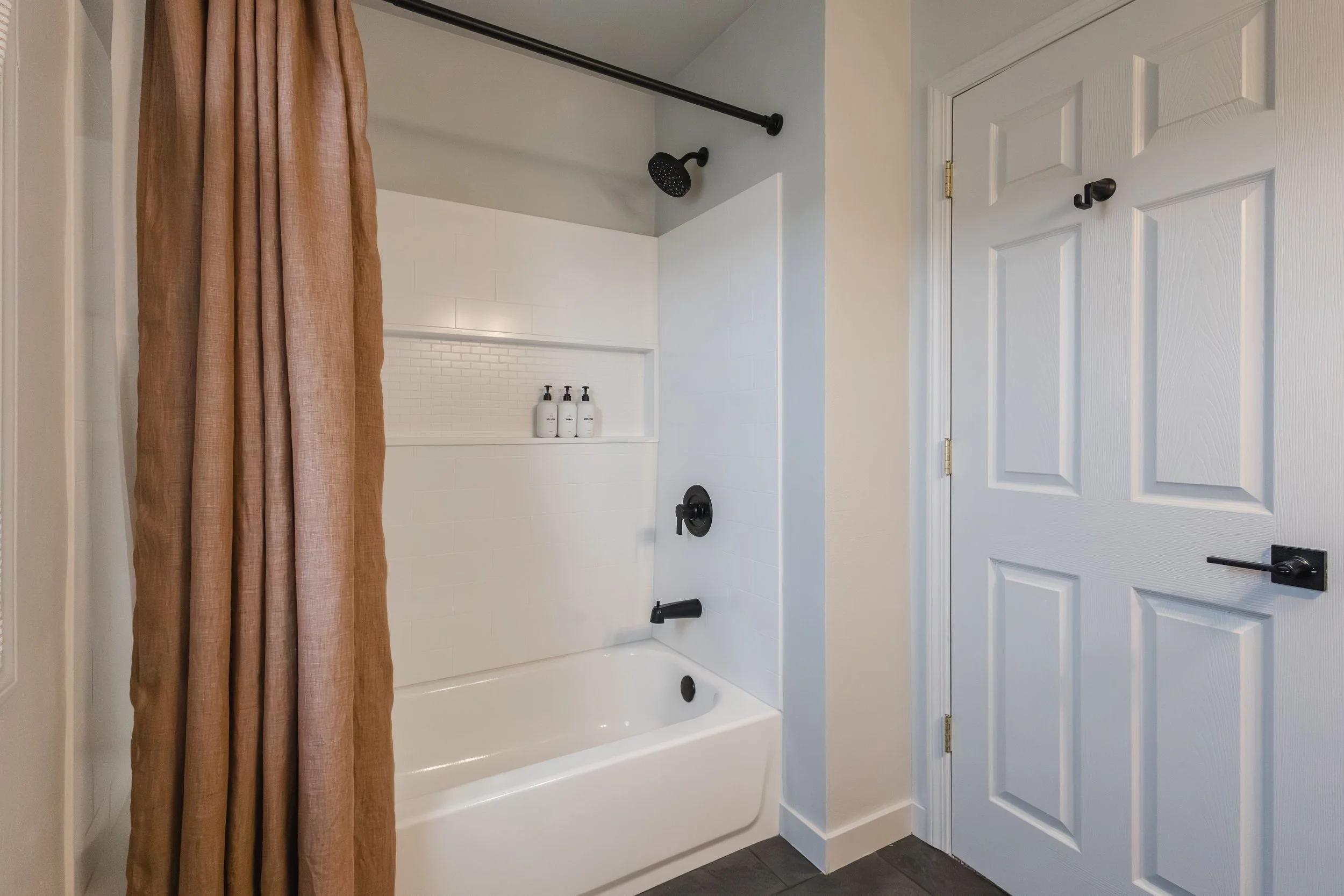 Bathroom shower with a white bathtub, a black showerhead, and a beige shower curtain. There are three white bottles on a white shelf inside the shower. The bathroom has a white door with black hardware and dark flooring.