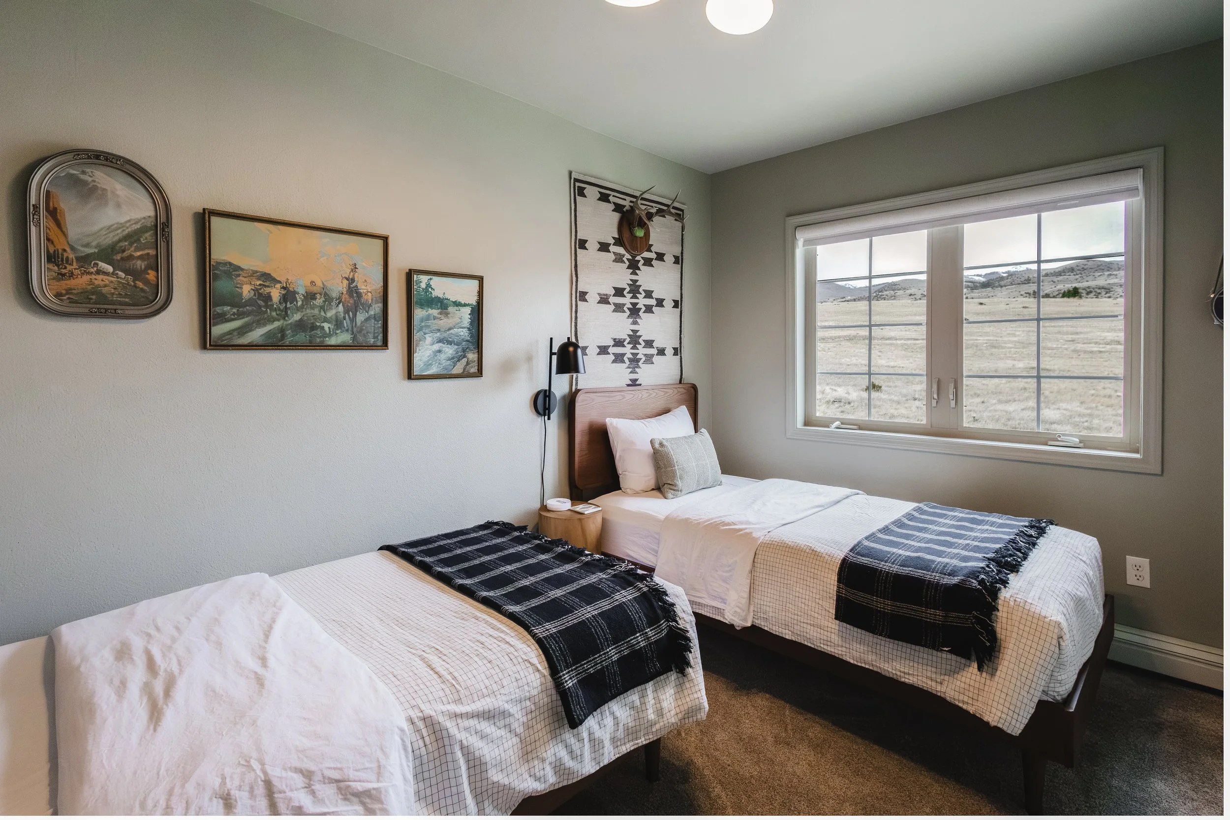 A bedroom with three beds, each with a blanket at the foot, pillows, and nightstand. A large window shows an outdoor landscape of hills and open land. Wall decorations include a woven textile and framed artwork, with a mounted antler on the wall.