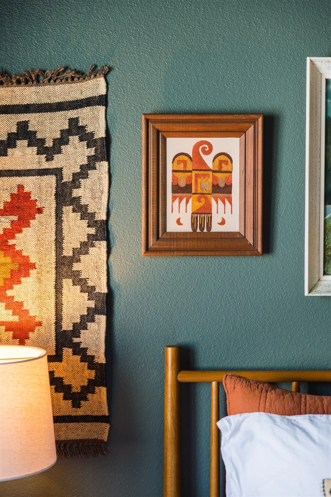 Decorative wall art with Southwestern patterns on a dark green wall, next to a wooden bed frame with a rust-colored pillow and white bedsheet, partially visible in a bedroom.