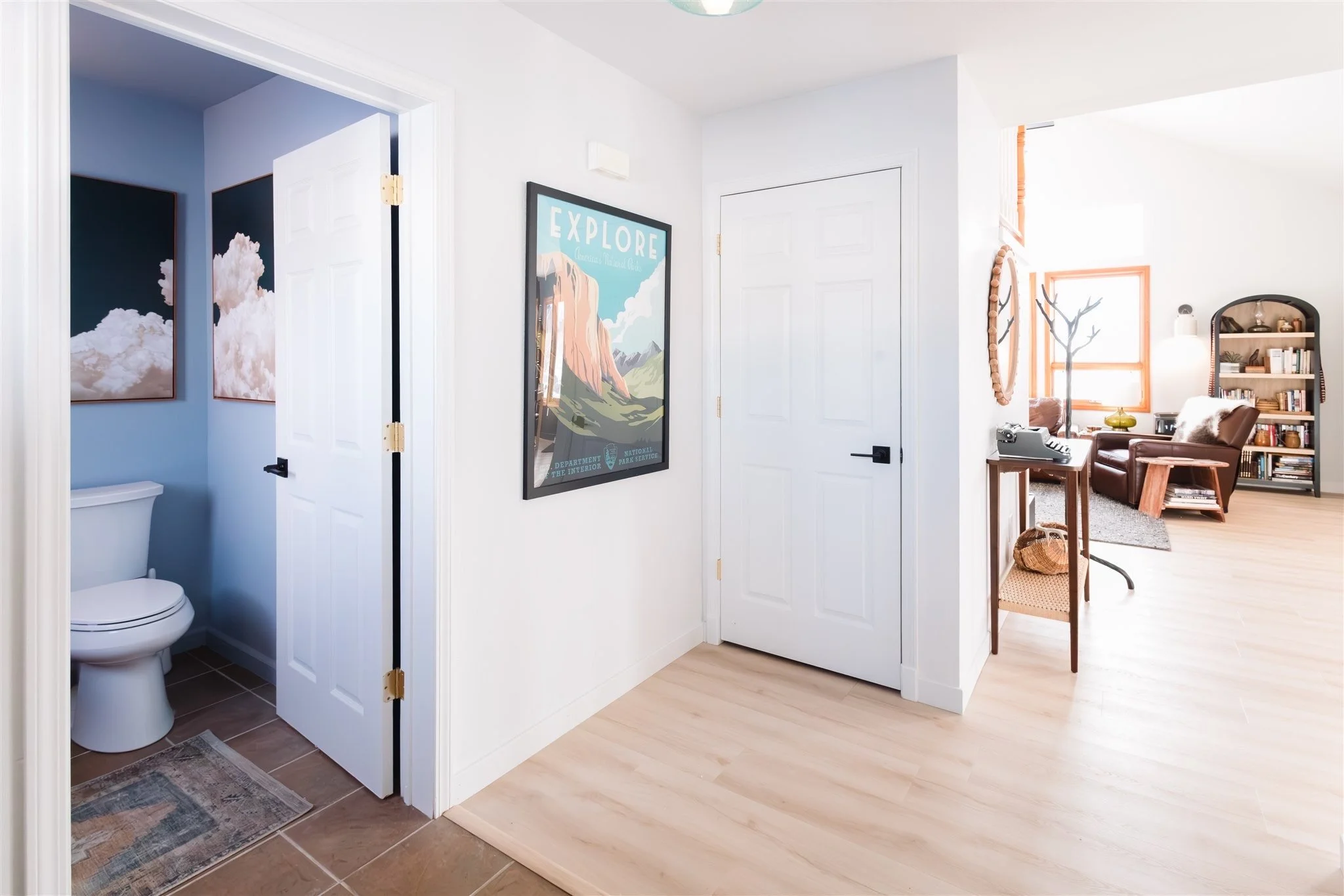 Interior of a home showing a hallway with an open door to a bathroom on the left, a decorative framed poster with the word 'EXPLORE' on the wall, and a view into a living room with a brown chair, bookshelf, plants, window with orange trim, and a lamp