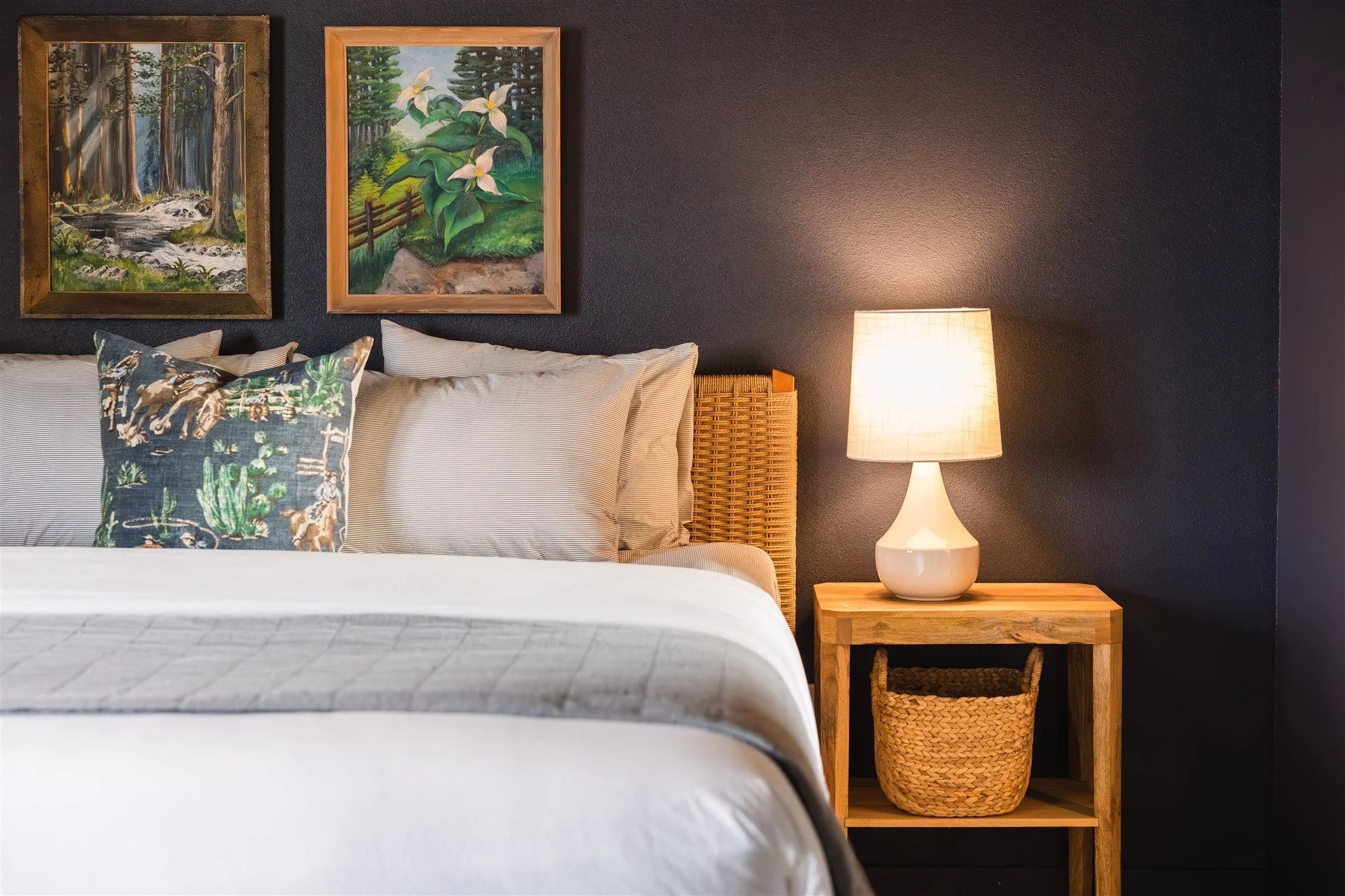 A bedroom with a dark accent wall, decorated with two framed landscape paintings. There is a bed with a white duvet and patterned pillows, a wooden nightstand with a woven basket underneath, a white table lamp, and soft lighting.