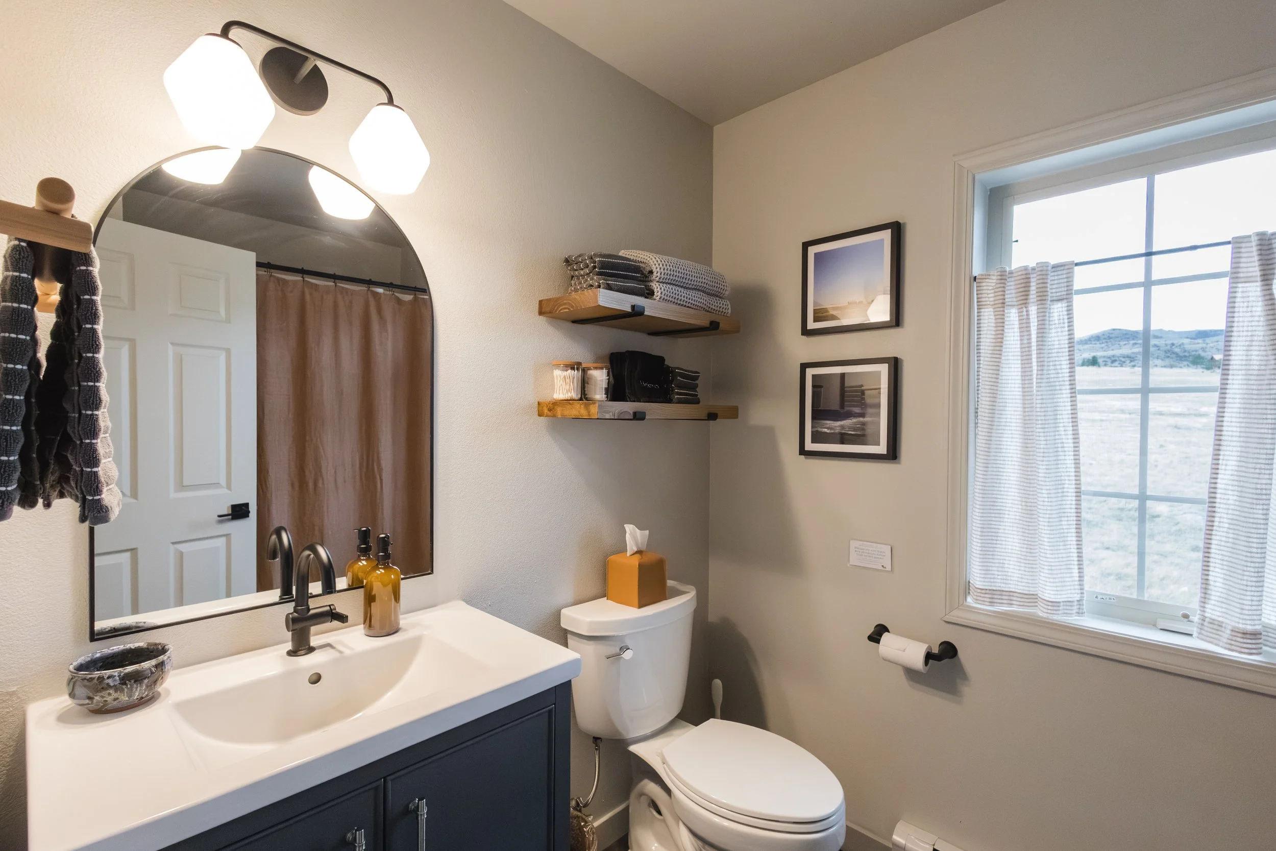 A bathroom with a white sink and dark cabinetry, a mirror above the sink, two soap dispensers, a toilet with a tissue box on top, a window with striped curtains, wall shelves with folded towels and bathroom supplies, and framed photos on the wall.