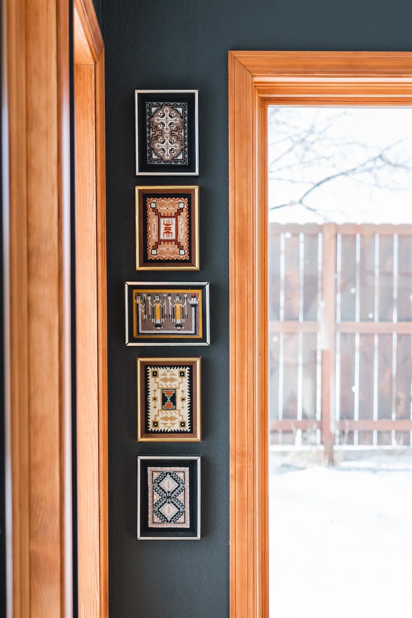A wall with five framed cross-stitch or embroidery artwork pieces arranged vertically, adjacent to a wooden doorframe and window with a view of an outdoor wooden fence.