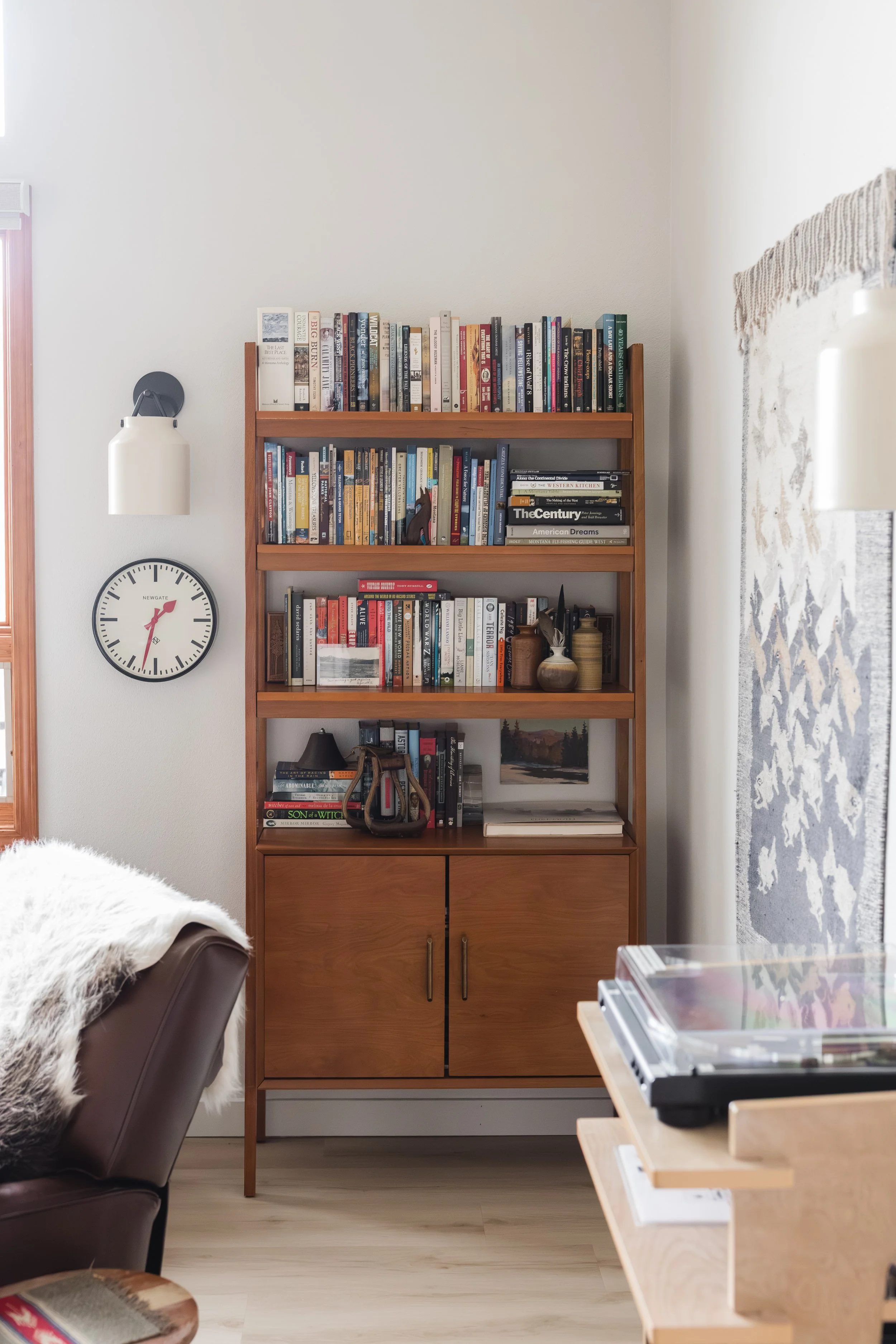 A wooden bookshelf filled with books, a wall clock, a wall light, and a part of a sofa with a throw blanket in a living room.