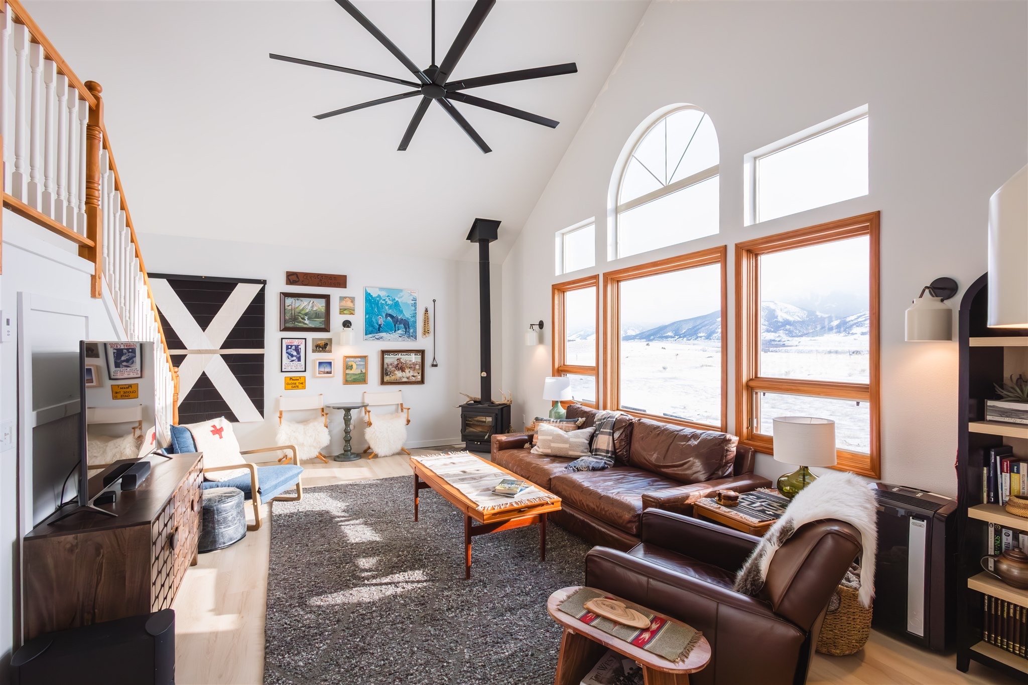 A living room with large windows showing snow-covered mountains outside. The room features a brown leather sofa, a coffee table, a black ceiling fan, a wood stove, and gallery wall art including paintings and photographs. 