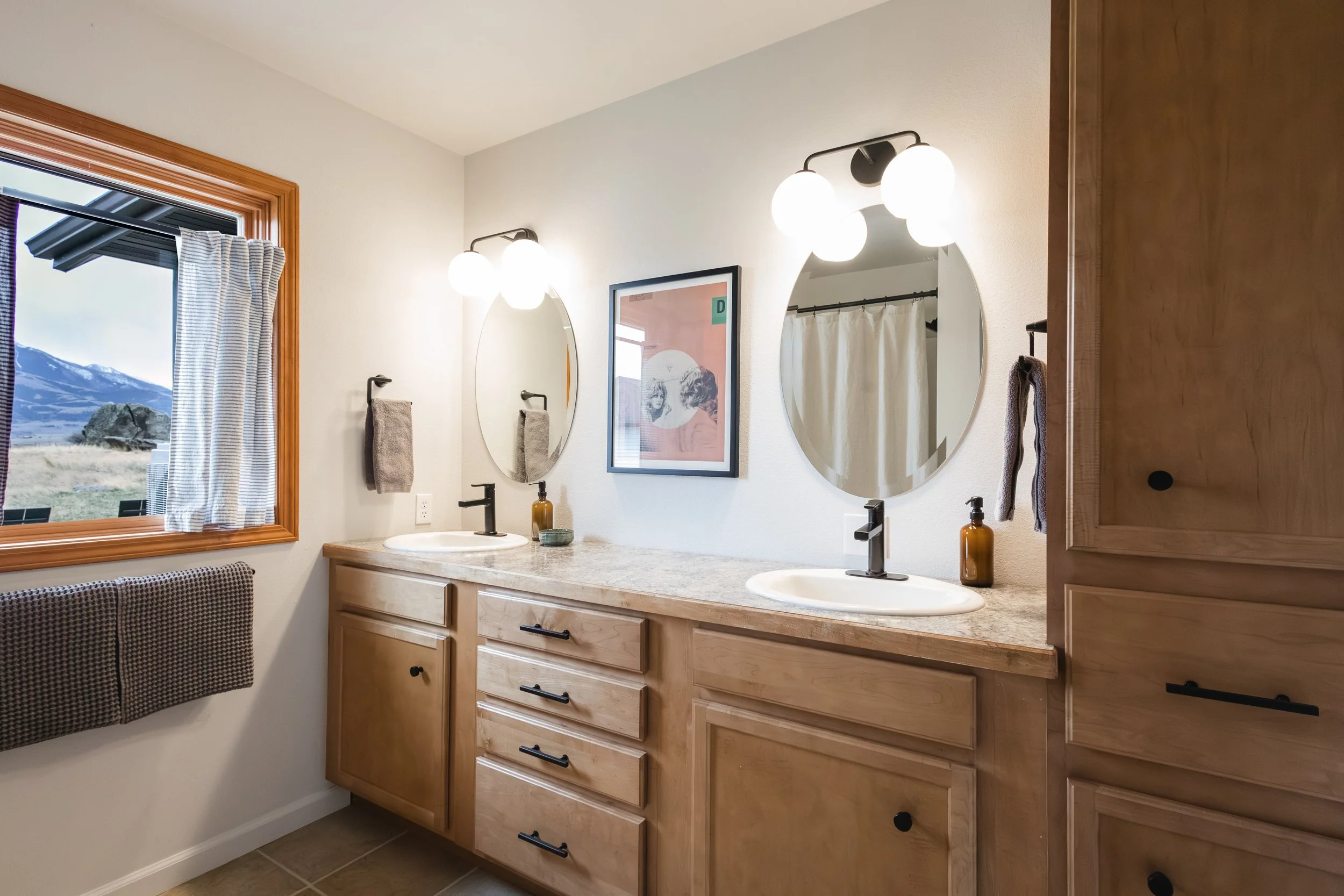A bathroom with a double vanity, two oval mirrors, black faucets, brown cabinets, and a window with a mountain view.