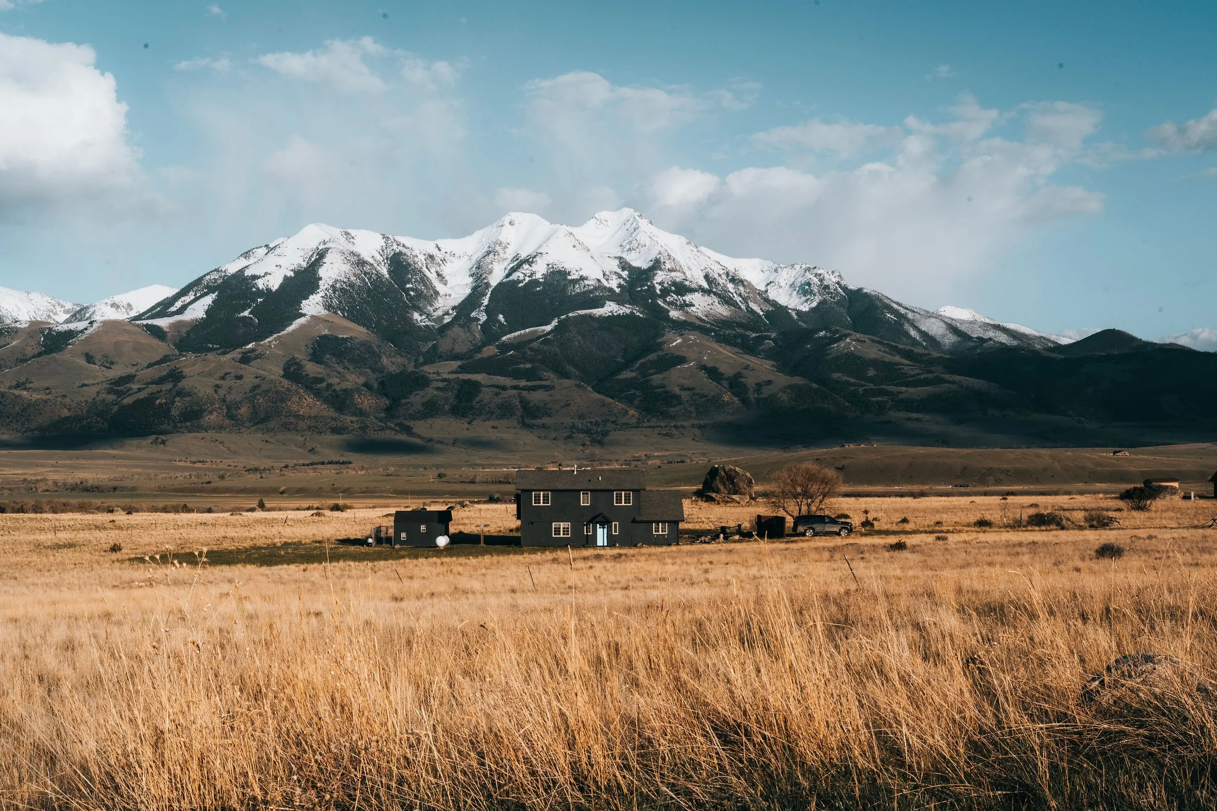 A landscape scene with tall native Montana prairie grass in the foreground, a dark vacation house, and snow-capped mountains under a partly cloudy sky in the background.