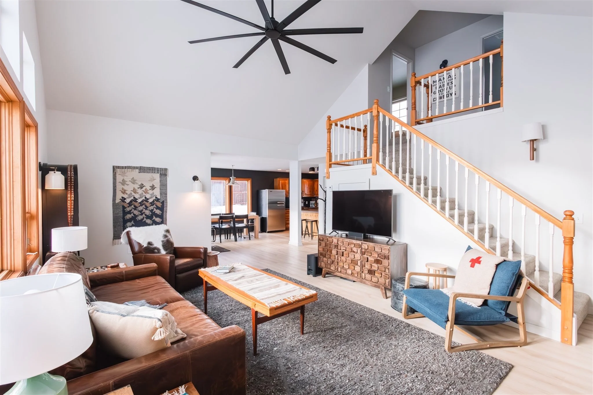 Living room with brown leather sofas,  lamps, a wooden coffee table, a blue armchair with a white pillow, a wall hanging with birds, a TV on a stand, staircase with wooden and white railing, and a view into the kitchen with dining table and appliance