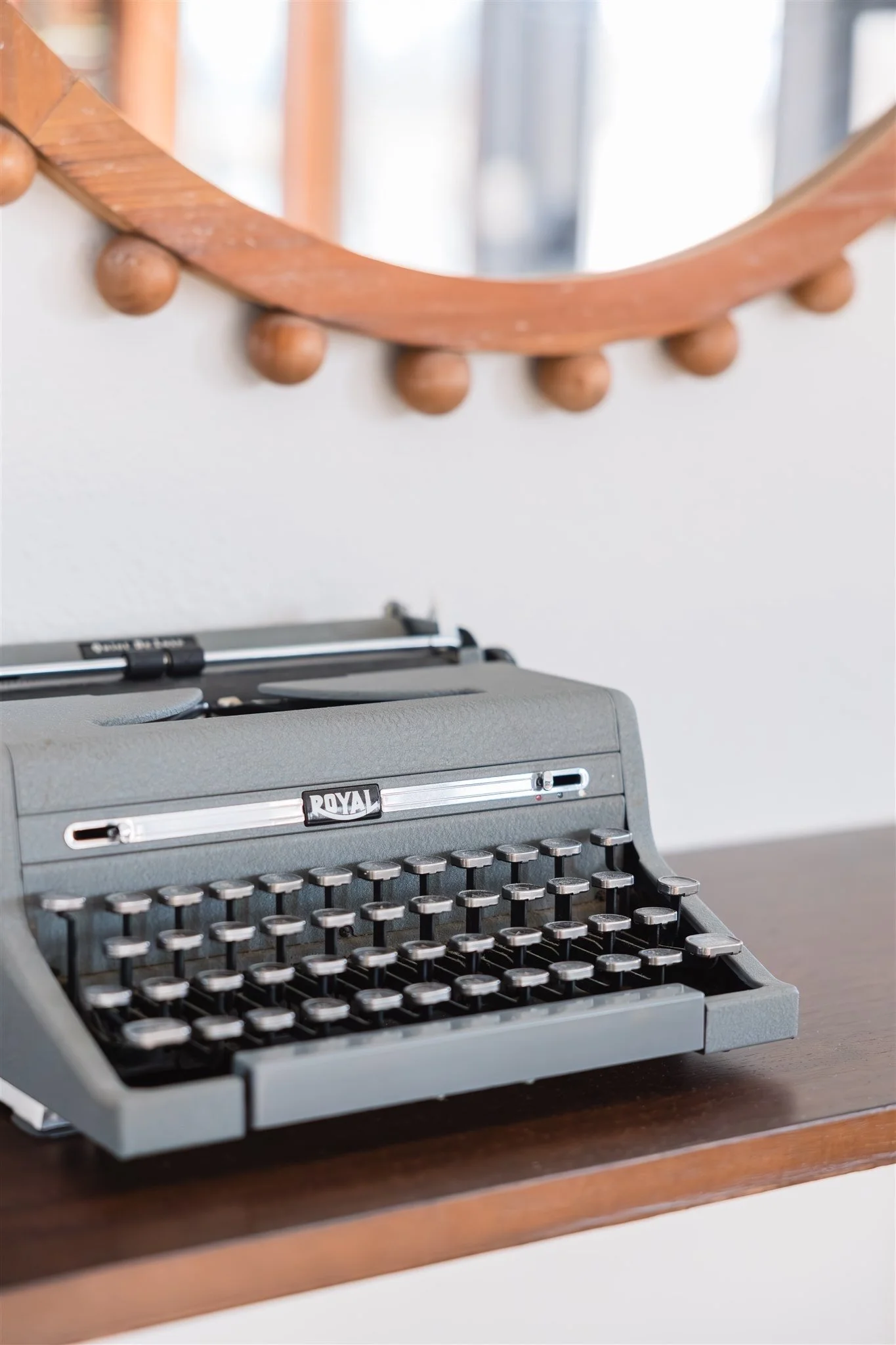 A vintage Royal manual typewriter sitting on a wooden desk with a decorative mirror with wooden frame hanging on the wall behind it.
