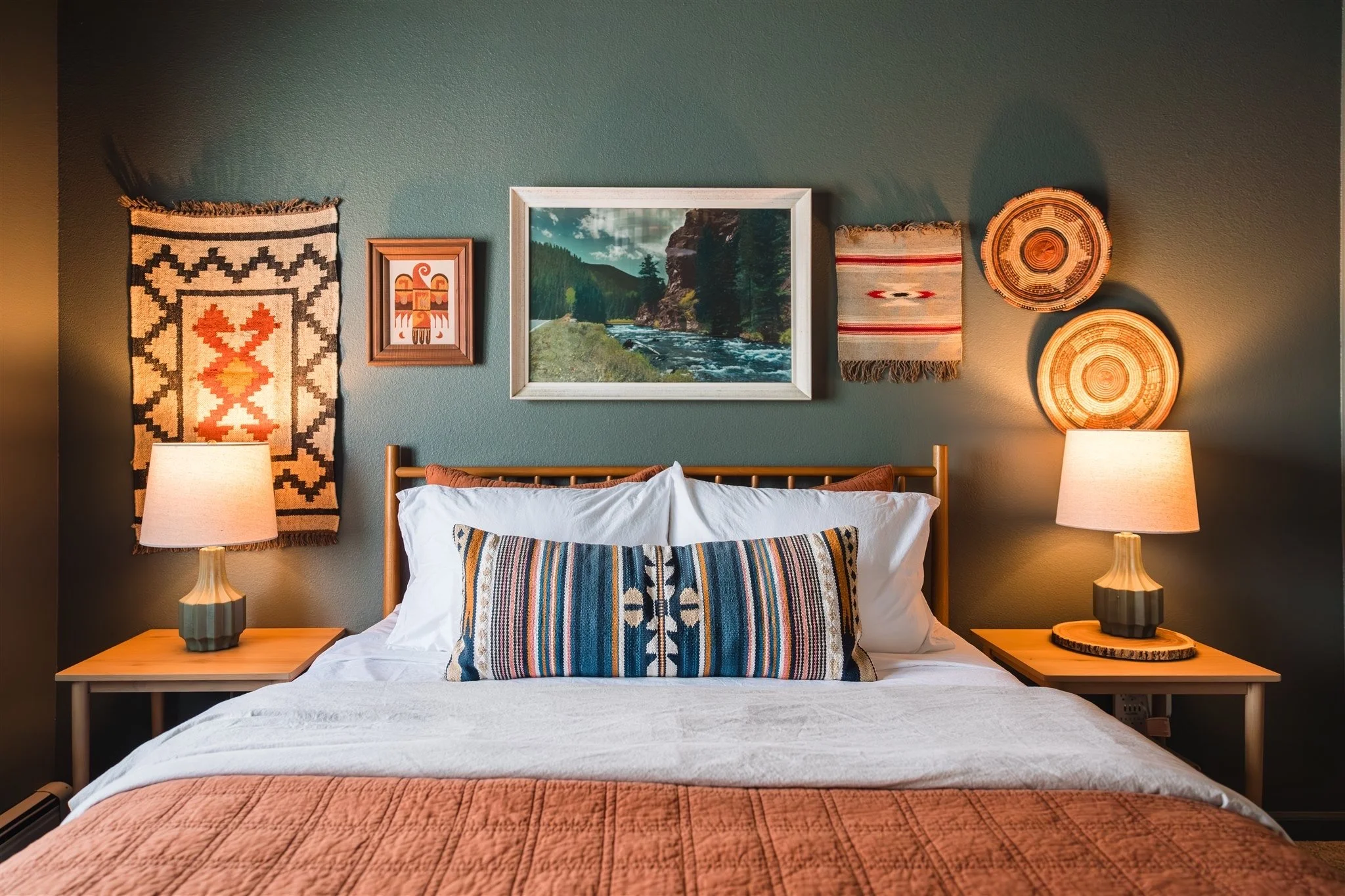 A bedroom with a bed, a turquoise wall decorated with artwork, two bedside tables with lamps, and a quilted bedspread.